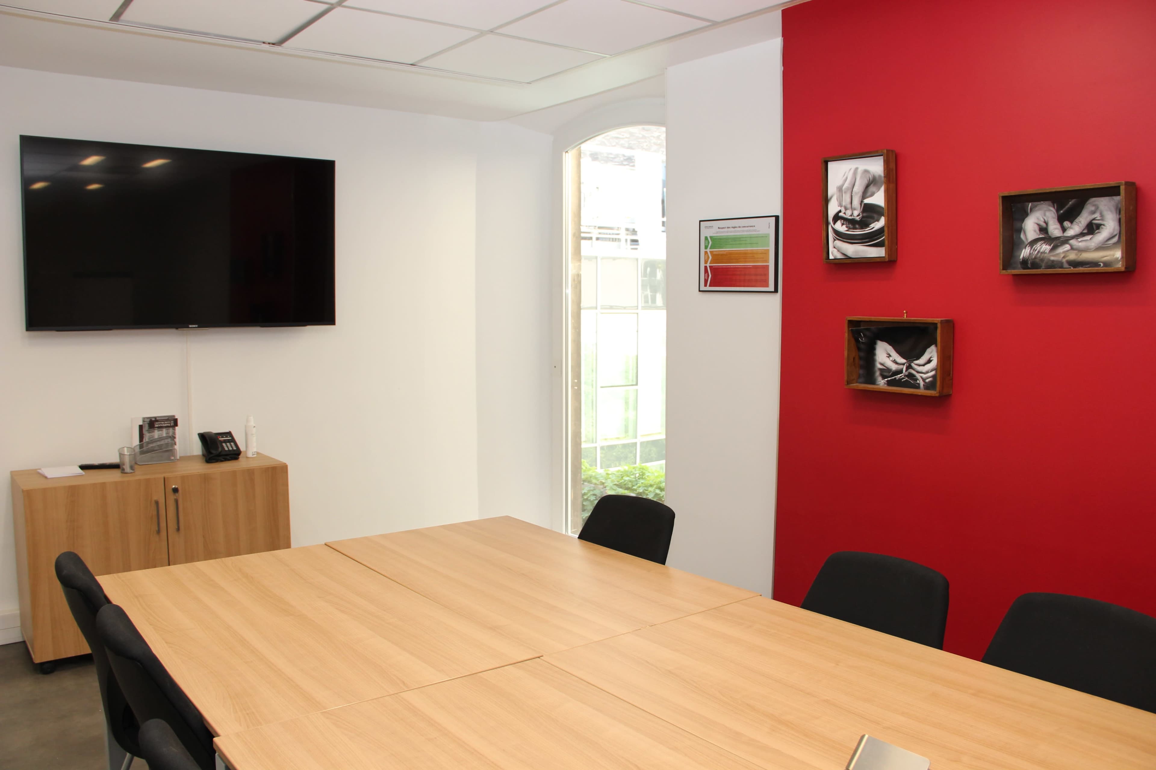 A conference room featuring a large table, black chairs, a wall-mounted television, and framed artwork on a red wall.