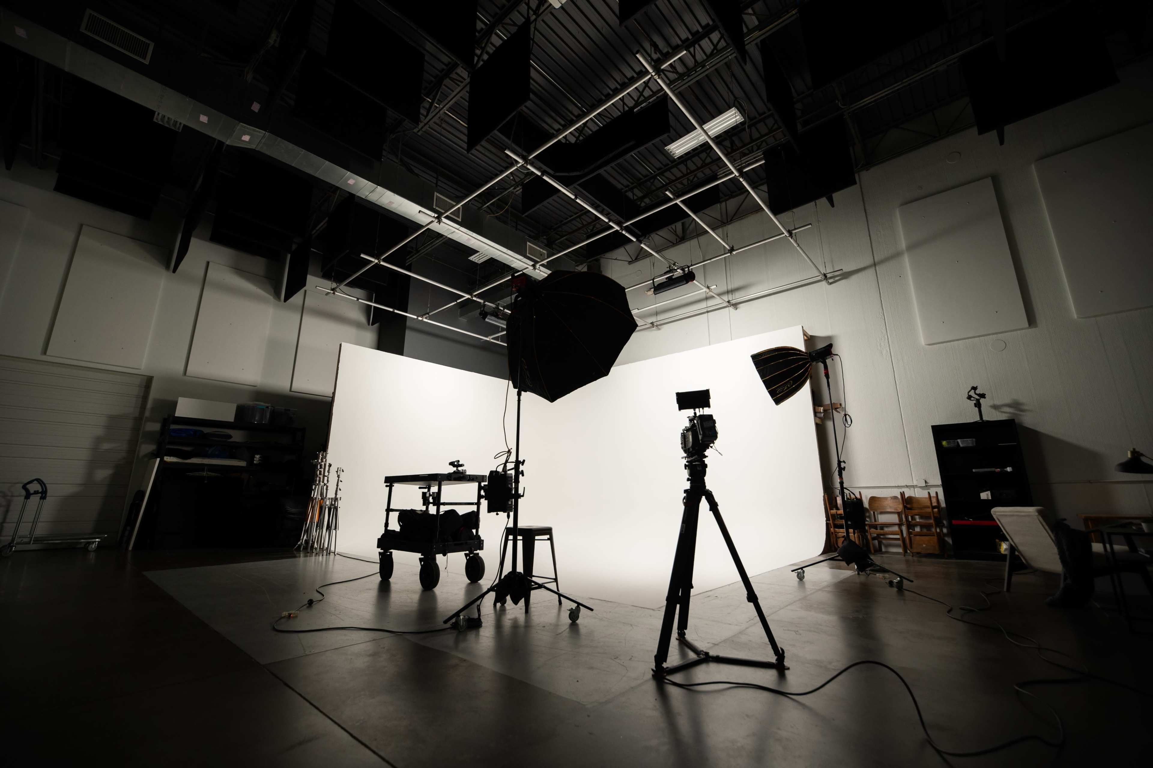 The image shows a photography or filming studio with a large white backdrop, lighting equipment, and a camera tripod arranged on a concrete floor.