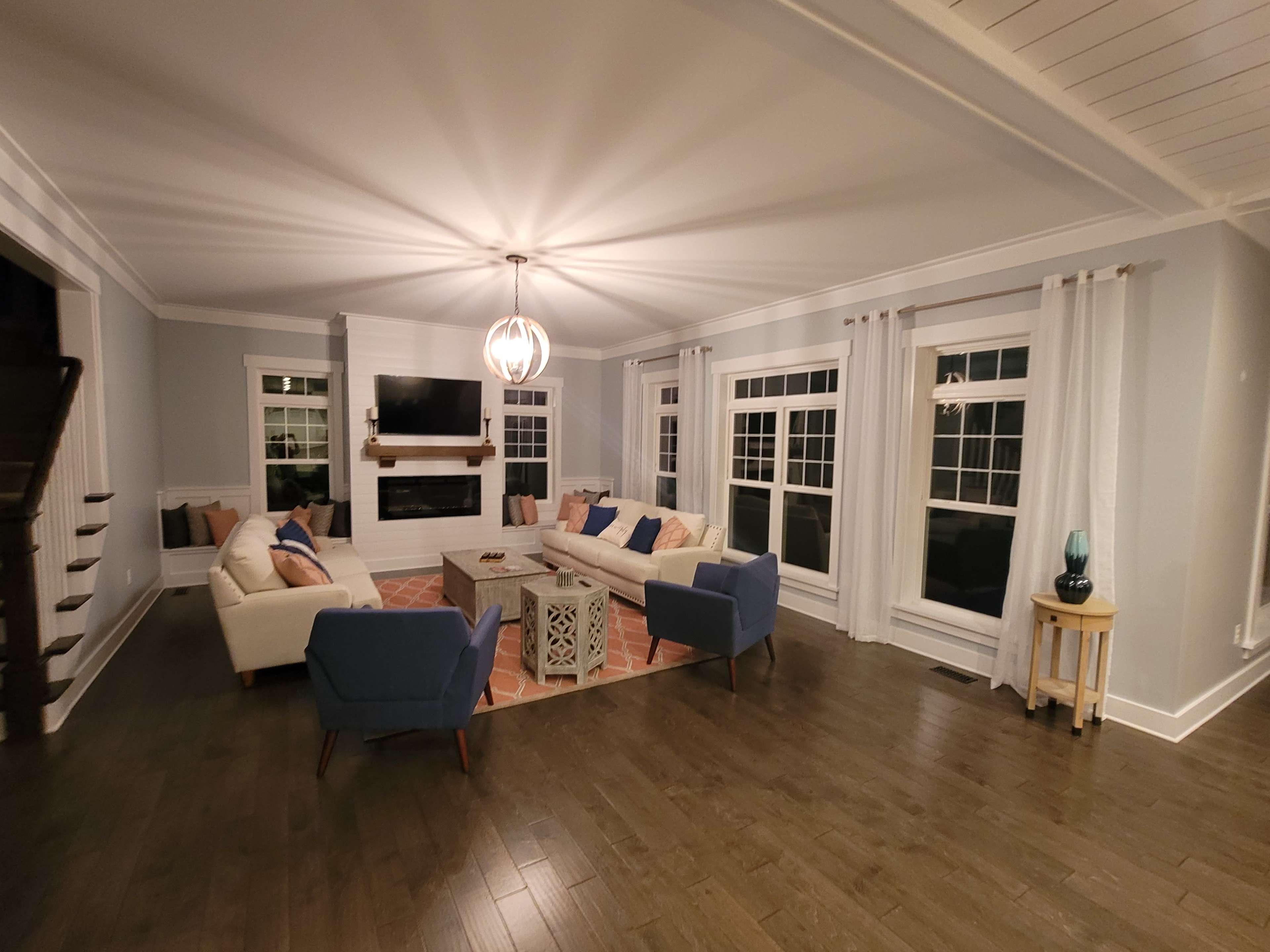 A spacious living room features a central coffee table surrounded by a sectional sofa and two blue armchairs, with large windows allowing natural light to enter.