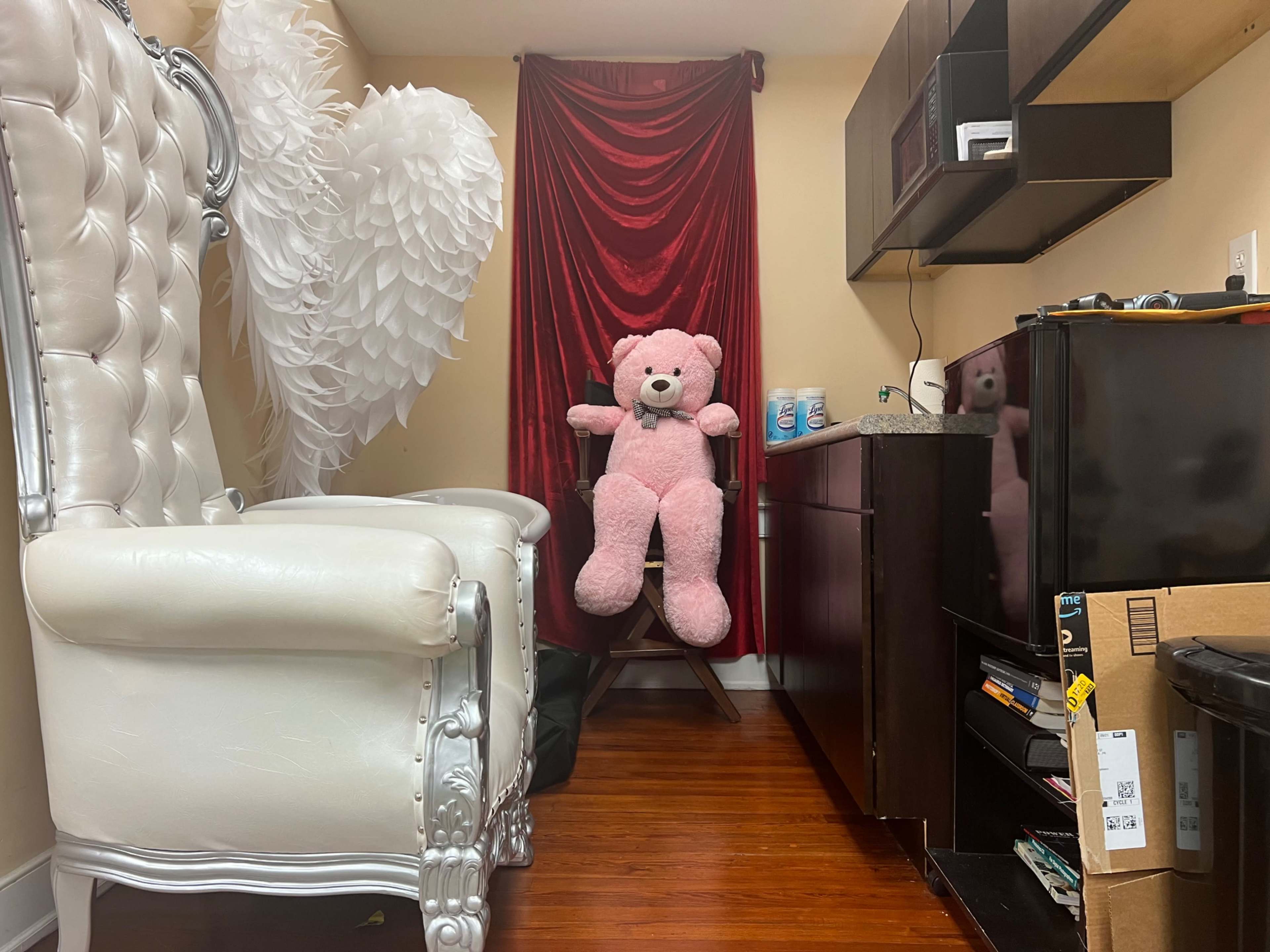The image shows a small room with a large white chair, a pink teddy bear sitting on a stool, and a red curtain in the background.
