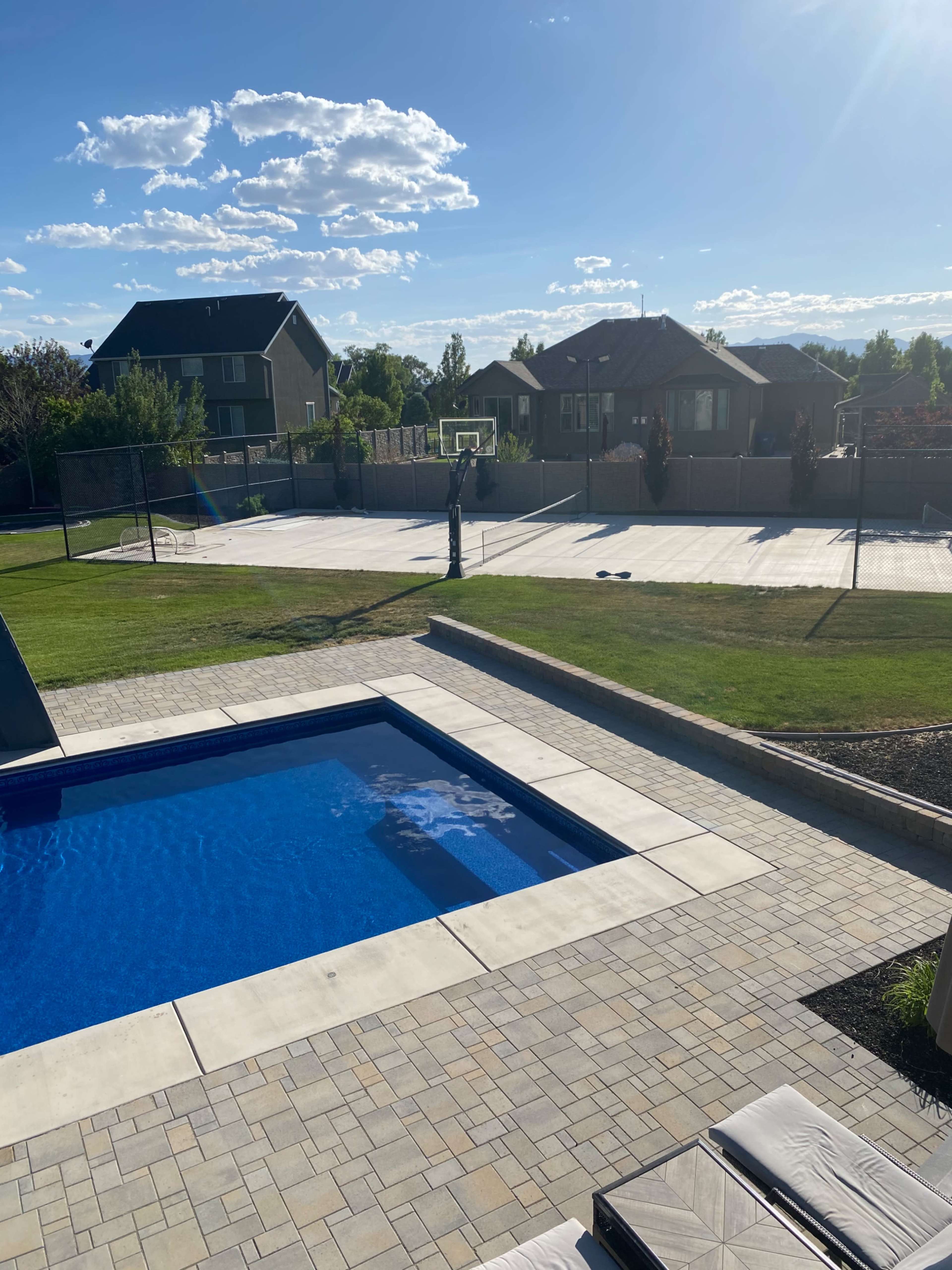 A person stands by a basketball hoop in a backyard with a pool and a well-maintained lawn.