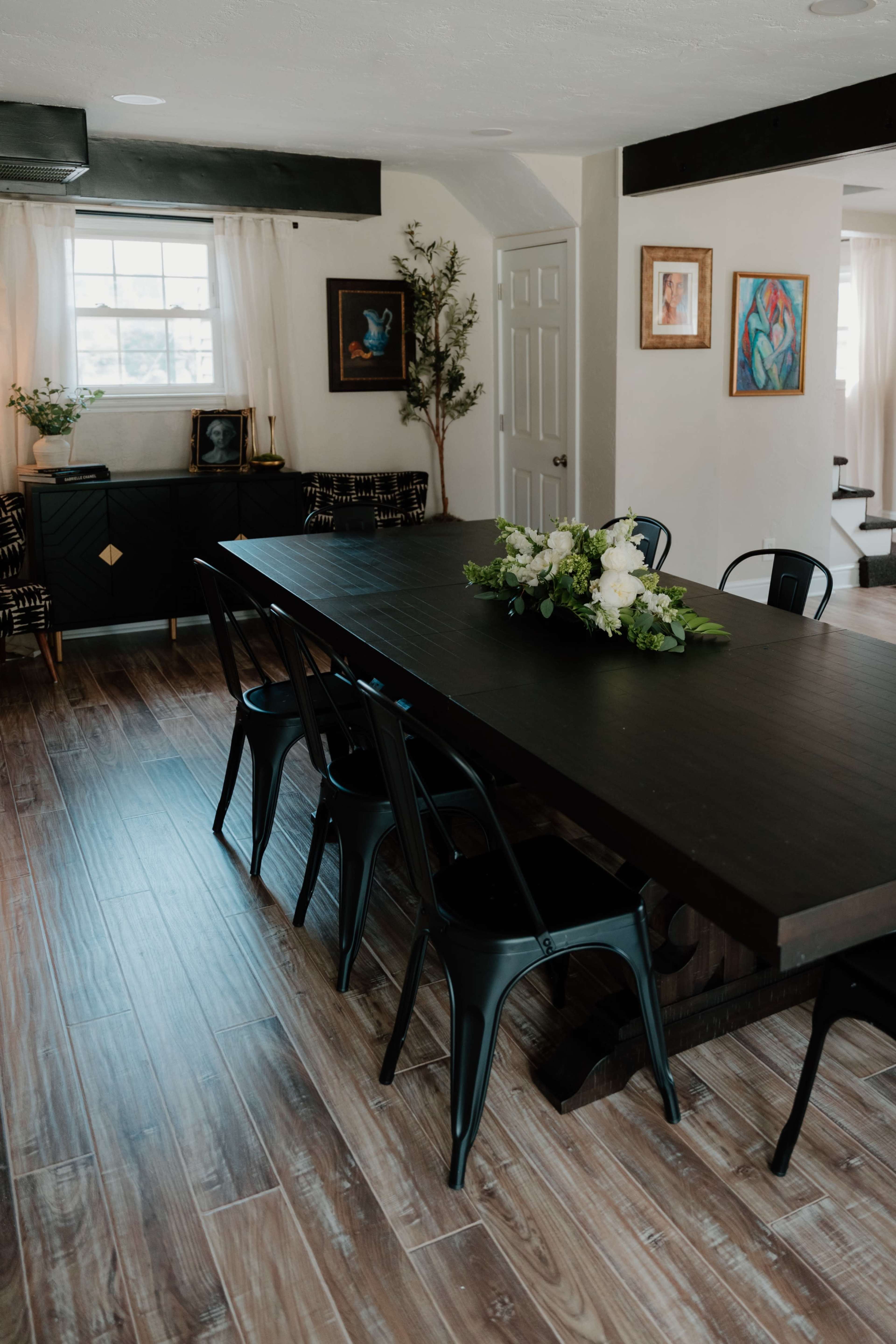 A long black dining table with metal chairs is set in a bright room featuring artwork and greenery.