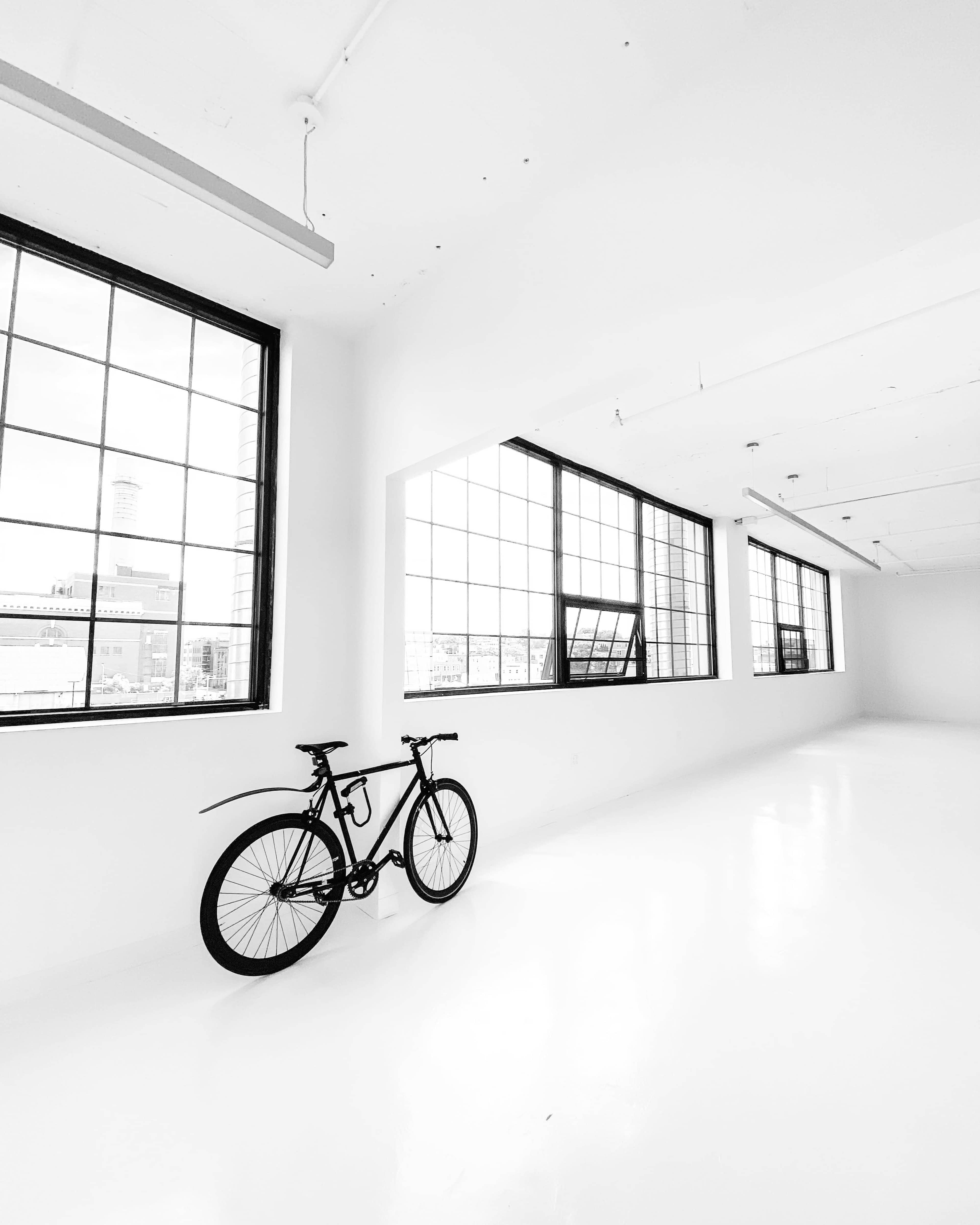A black bicycle leans against a white wall in a spacious, well-lit room with large windows.