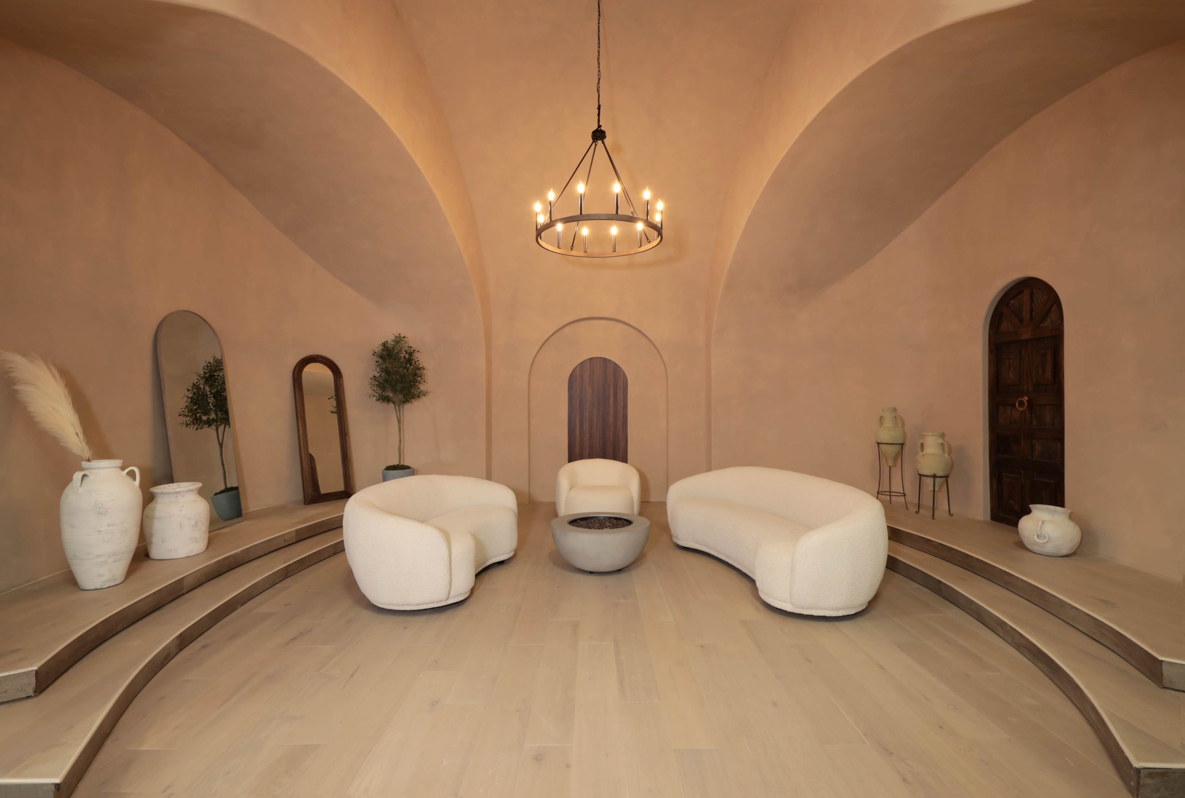 A spacious, minimalist interior with curved seating arrangements, a central table, and decorative elements such as mirrors and pottery set against a softly lit, arching wall.