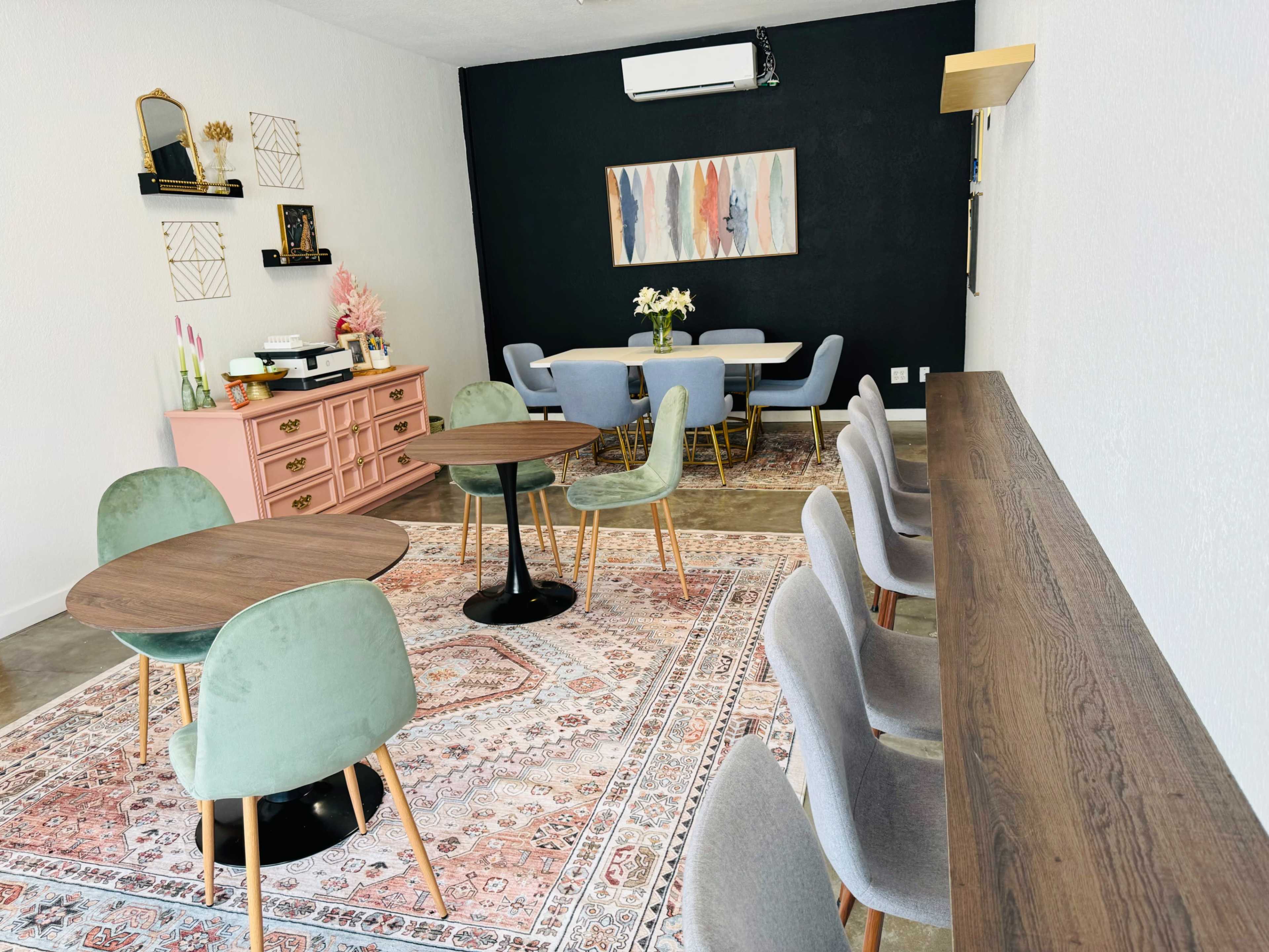 A modern interior space featuring a set of tables and chairs arranged around a decorative rug, with a pink dresser and a wall-mounted artwork in the background.