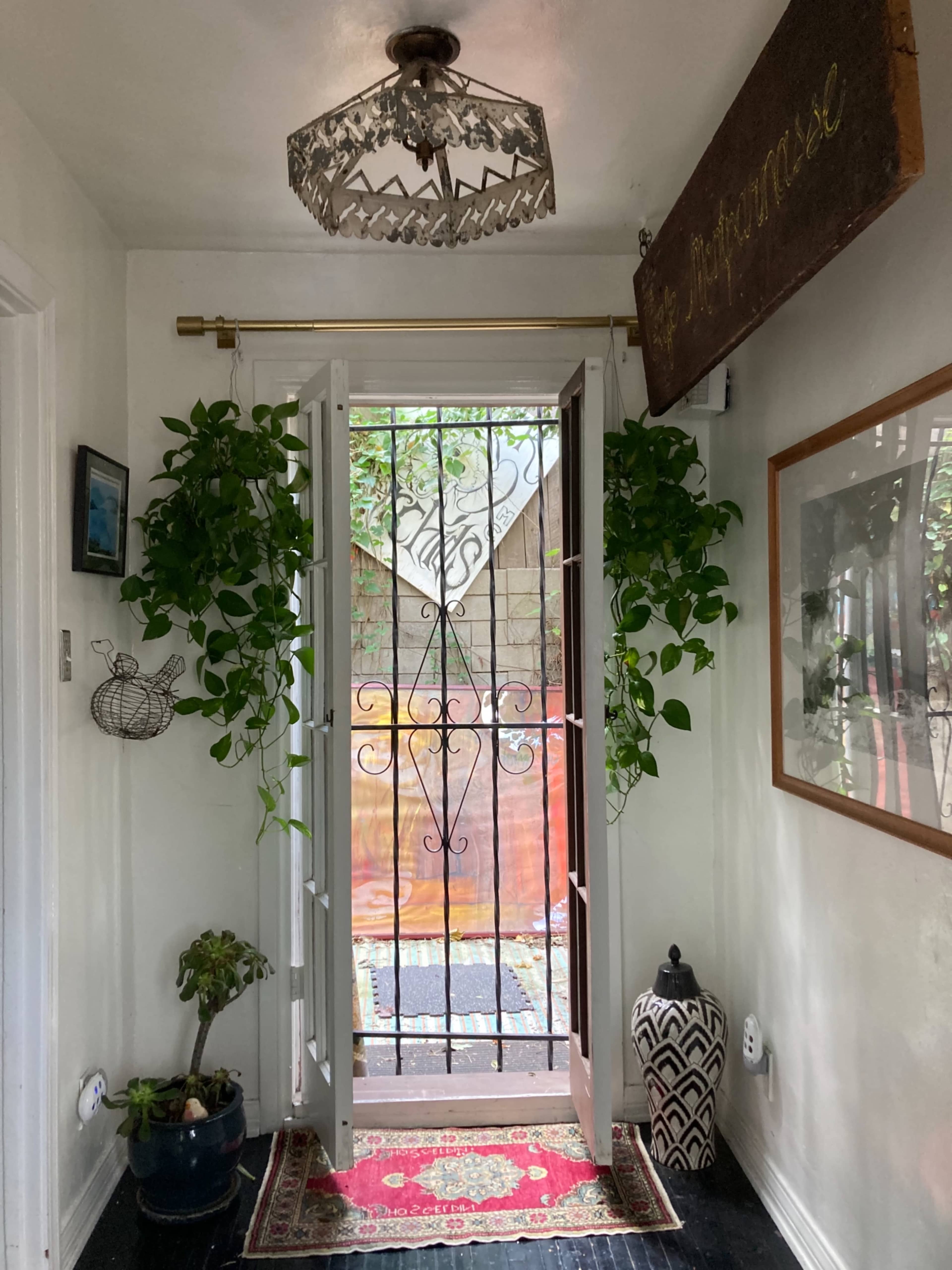 A bright hallway features an open door with ornate ironwork, flanked by potted plants and a patterned rug.