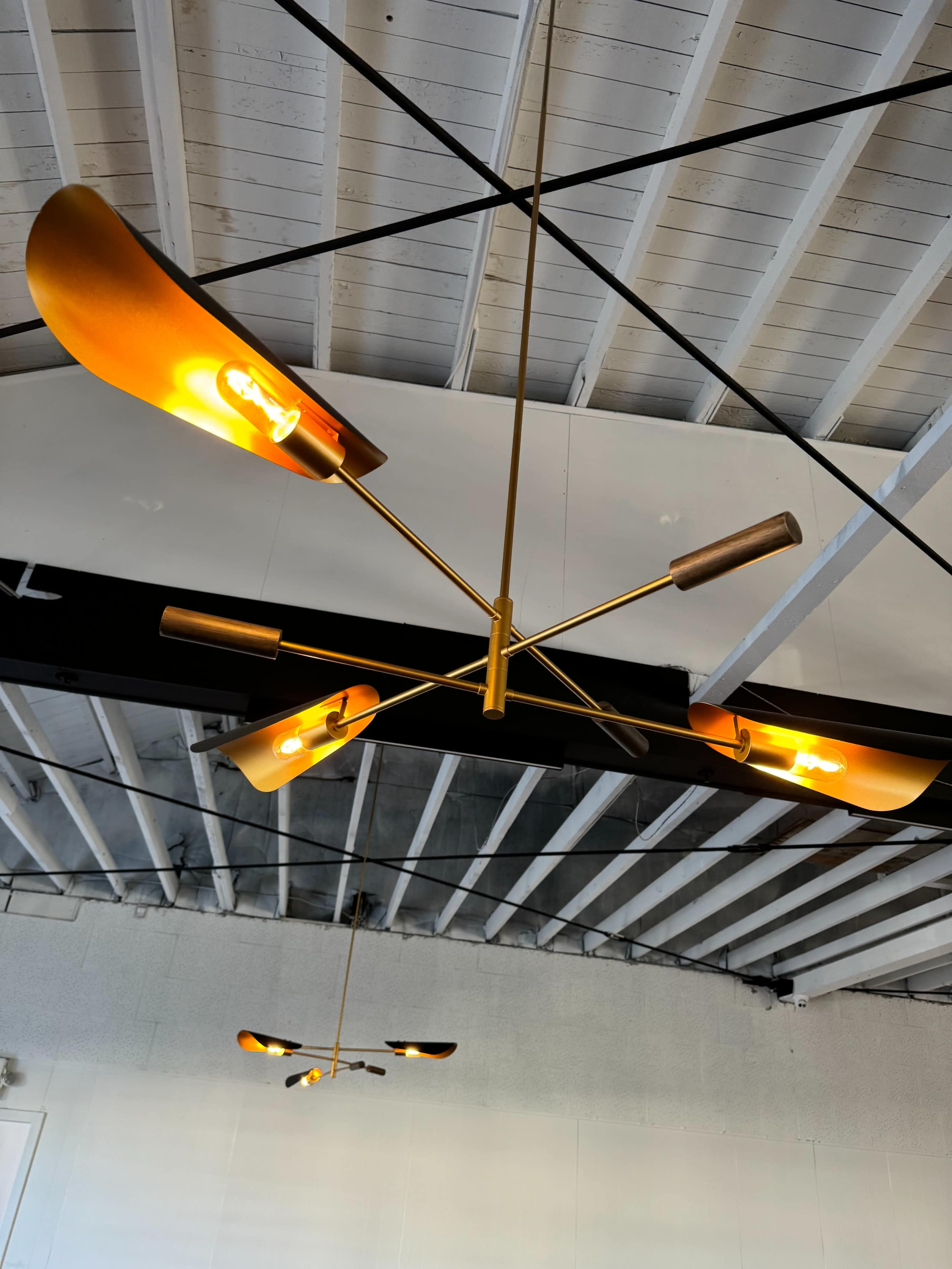 The image depicts a modern ceiling light fixture with four arms and curved shades, suspended from a black-beamed ceiling.