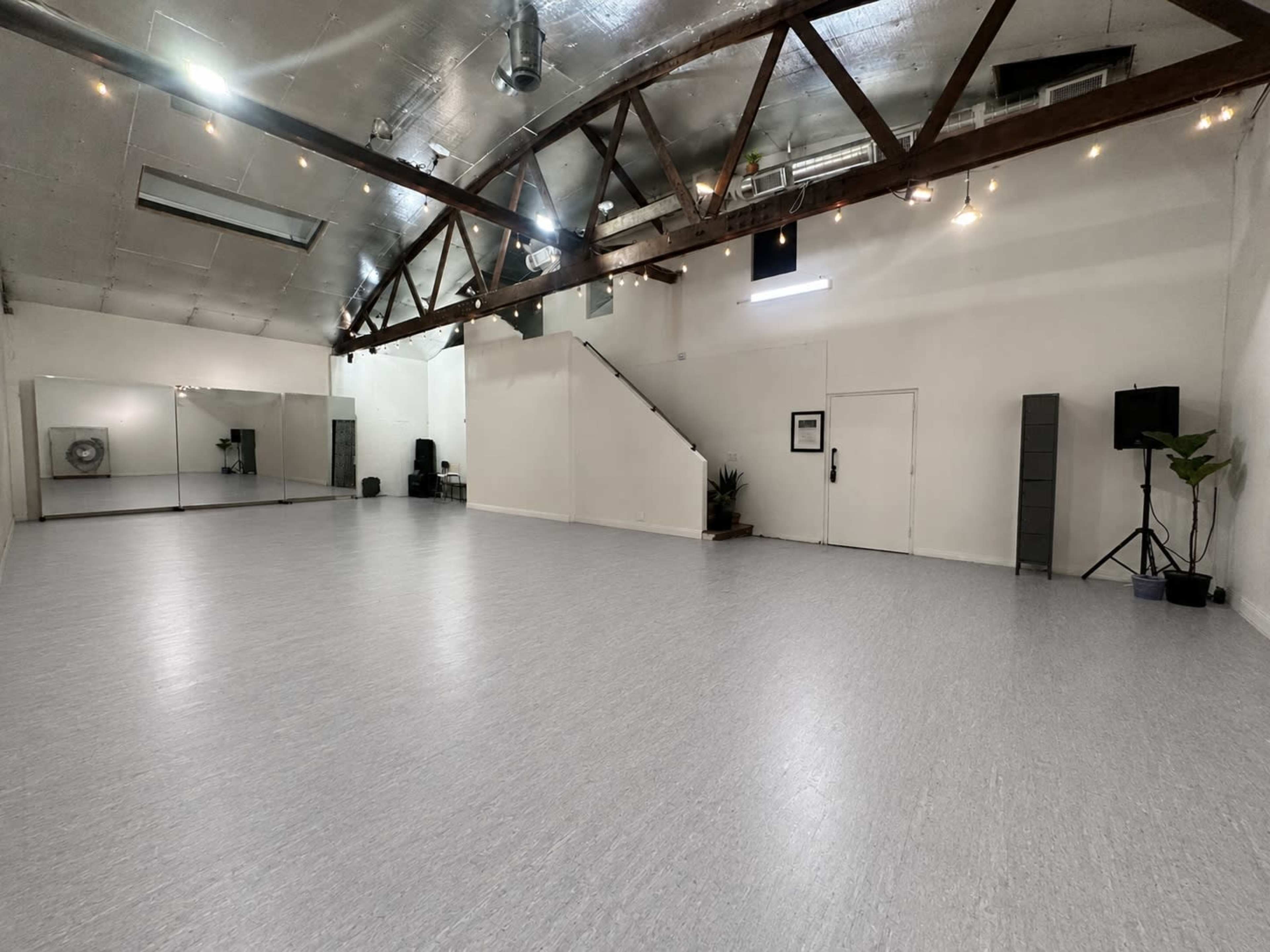 The image shows a spacious dance studio with high ceilings, wooden beams, a mirrored wall, and a staircase leading to an upper level.