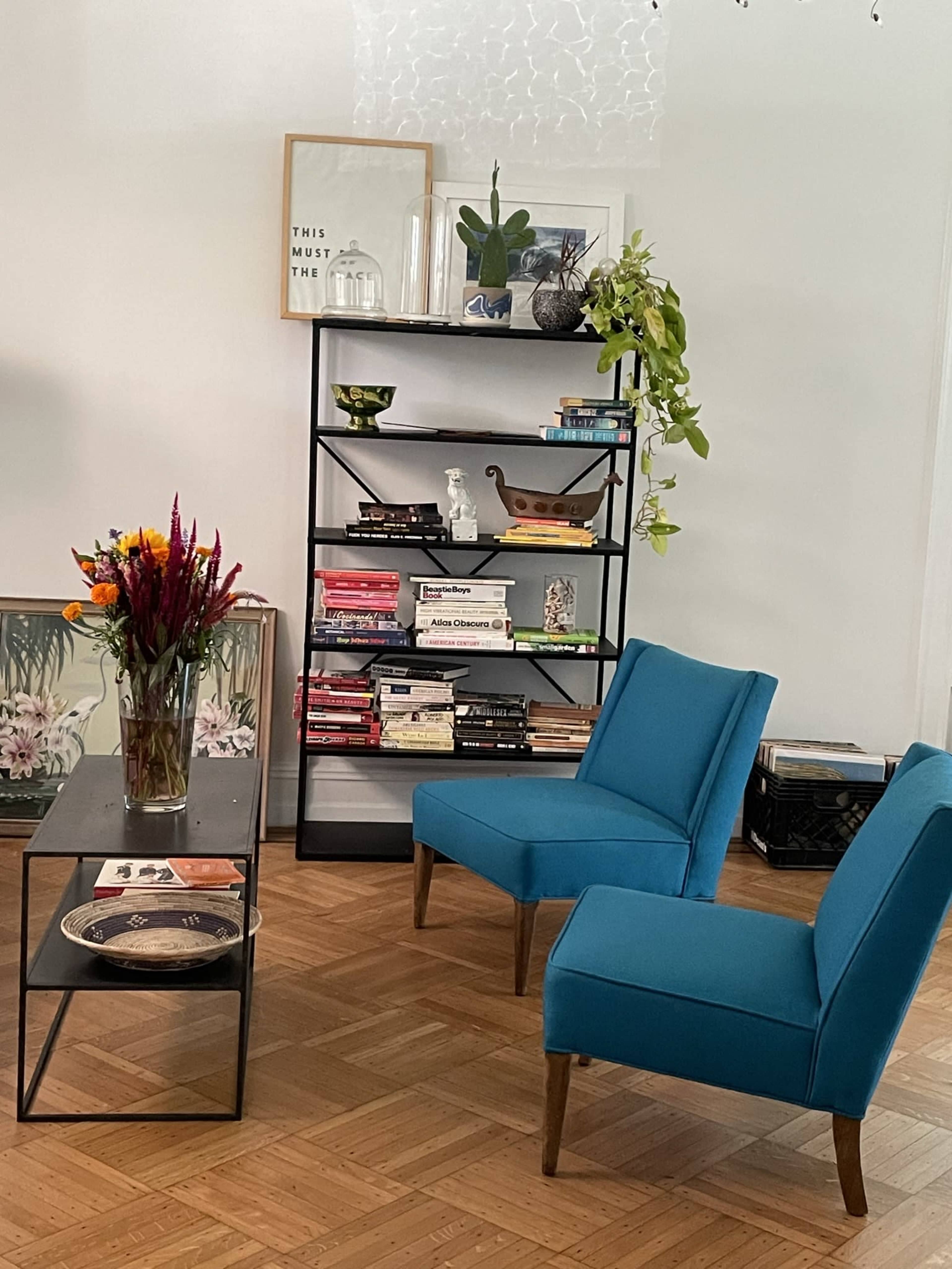 A cozy living room corner with two blue chairs, a black bookshelf filled with books, and a vase of flowers on a glass table.