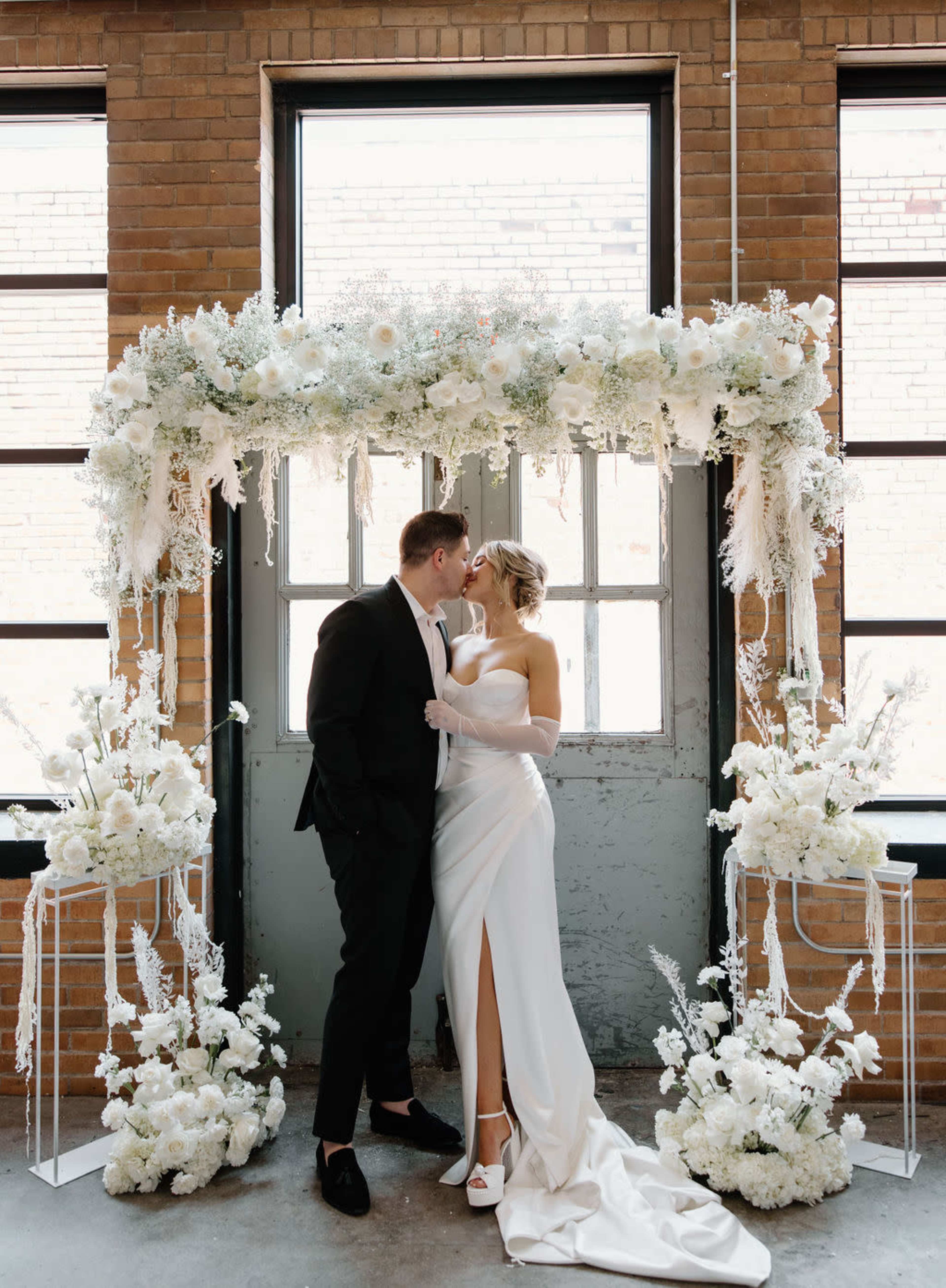 A couple stands in front of a floral arch, sharing a kiss in a venue with exposed brick walls and large windows.