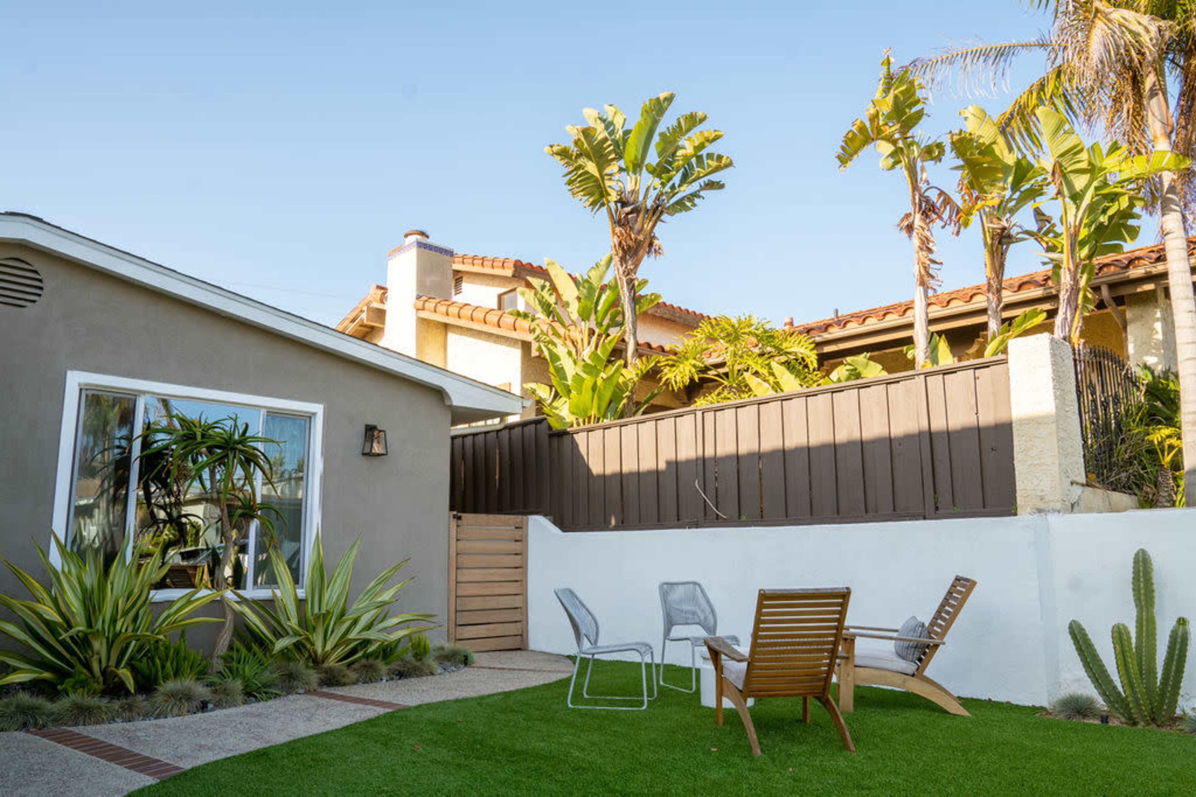 A modern backyard features a green lawn, a seating area with two chairs and a side table, surrounded by tall plants and a wooden fence.