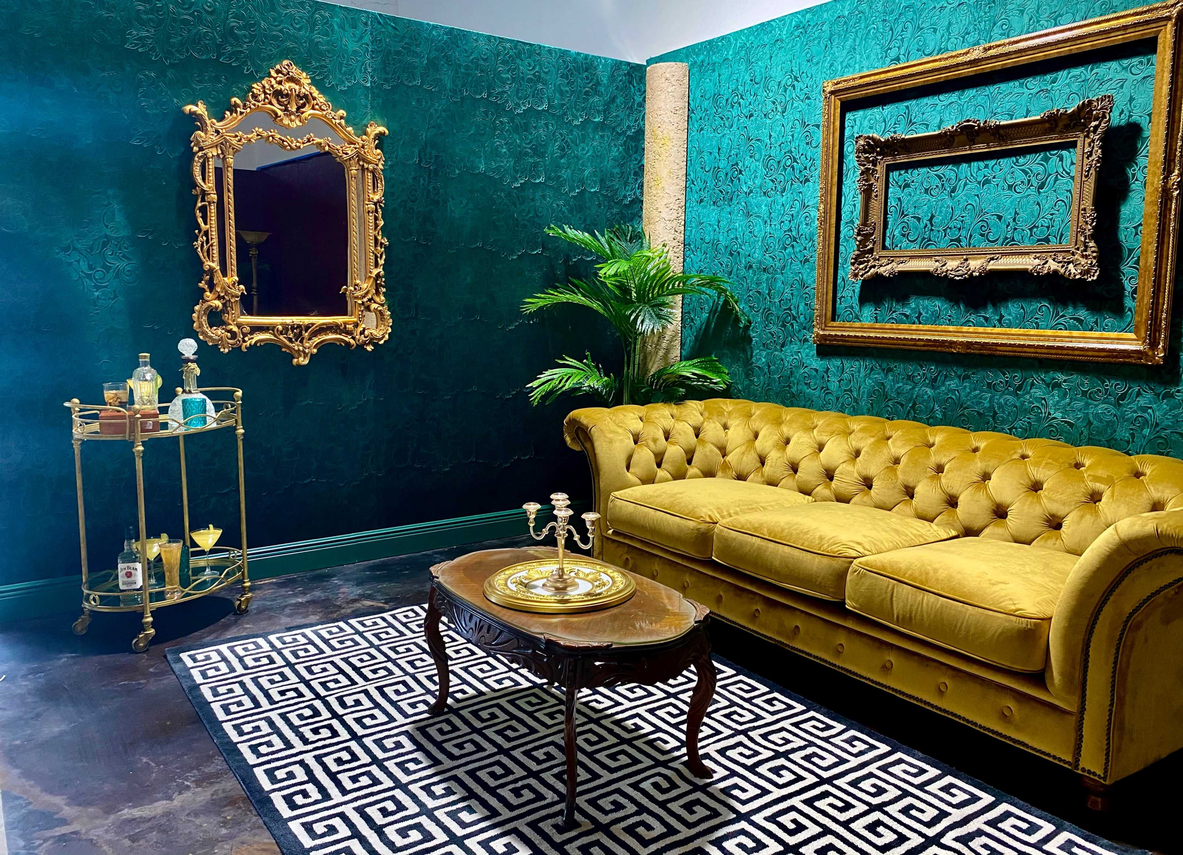 A vintage-inspired sitting area with a yellow tufted sofa, a decorative mirror, and a bar cart on a patterned rug against a teal wall.