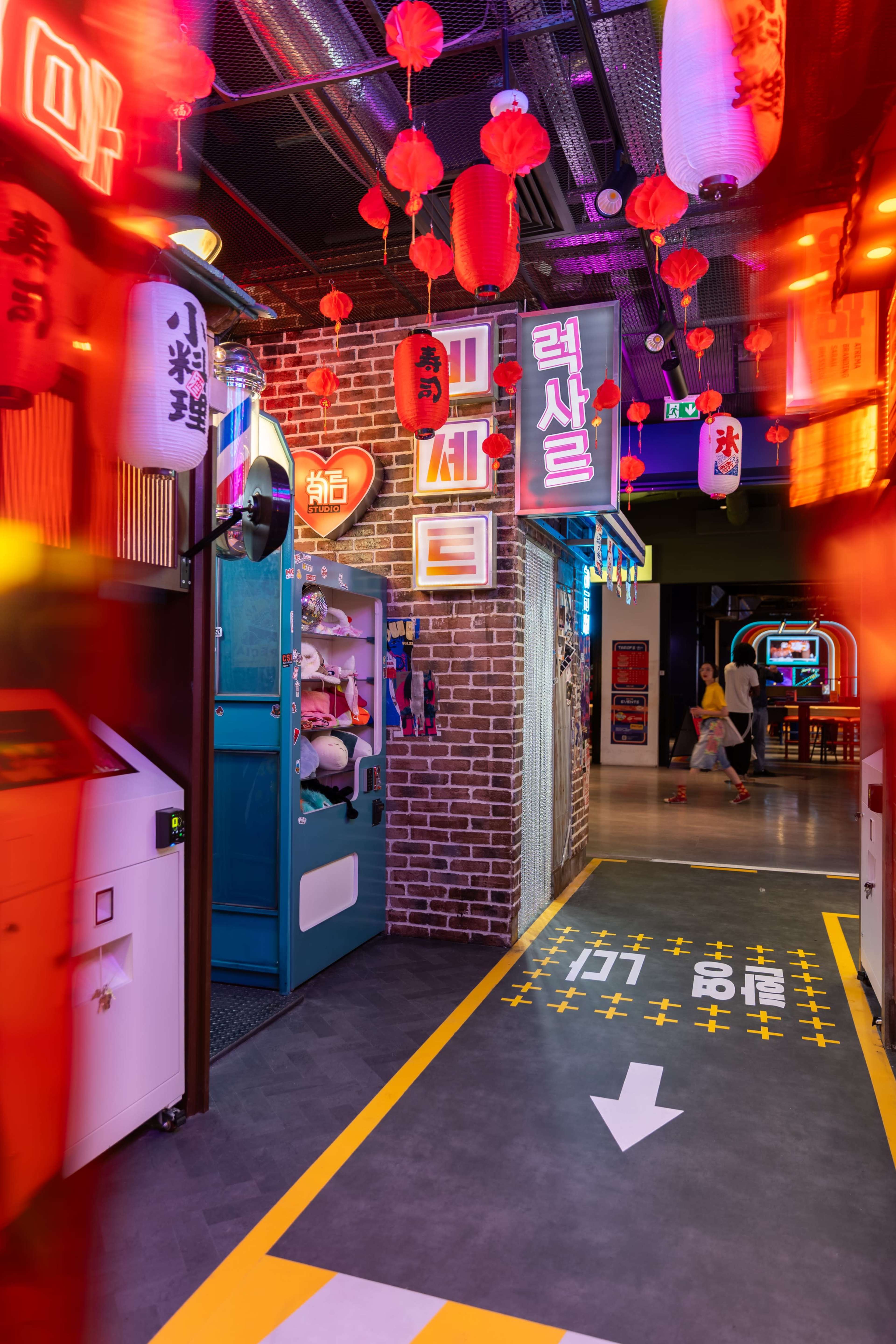 The image shows a vibrant indoor entertainment area featuring glowing signs, lanterns, and a pathway marked with arrows.