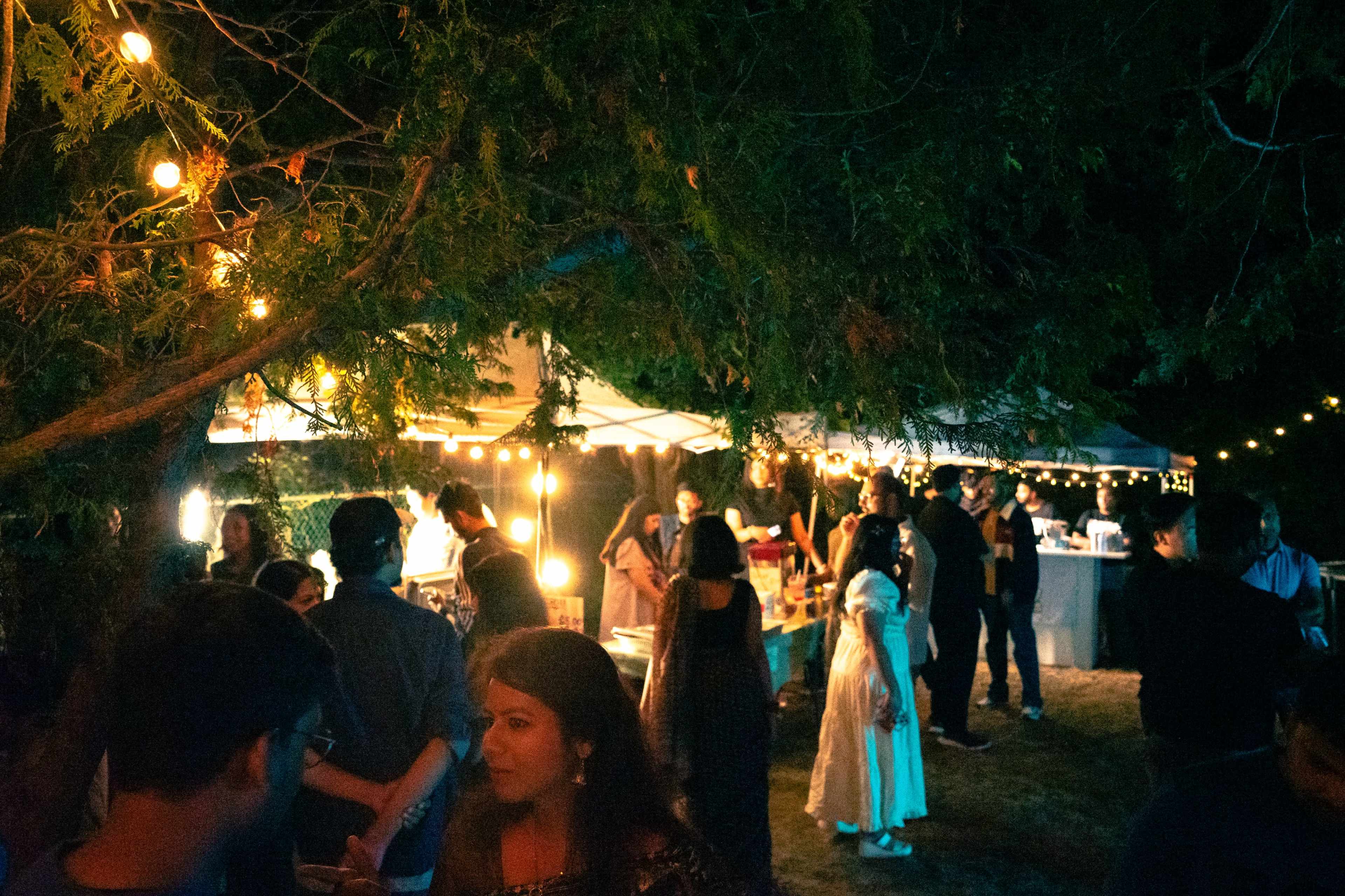 A lively outdoor gathering features illuminated tents and people socializing under string lights among trees.