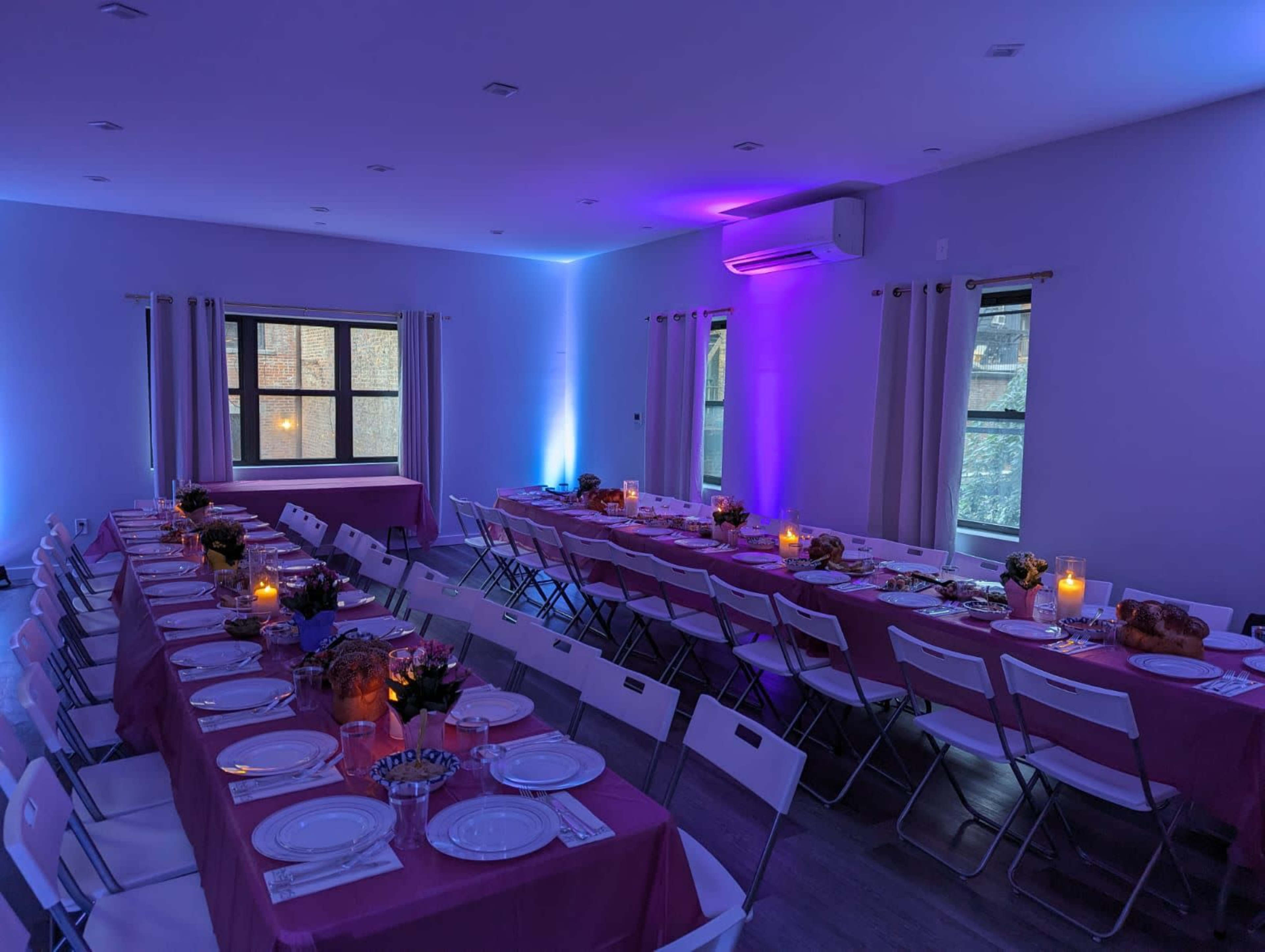 A brightly lit event space features multiple long tables set with plates and flowers, illuminated by purple lighting.