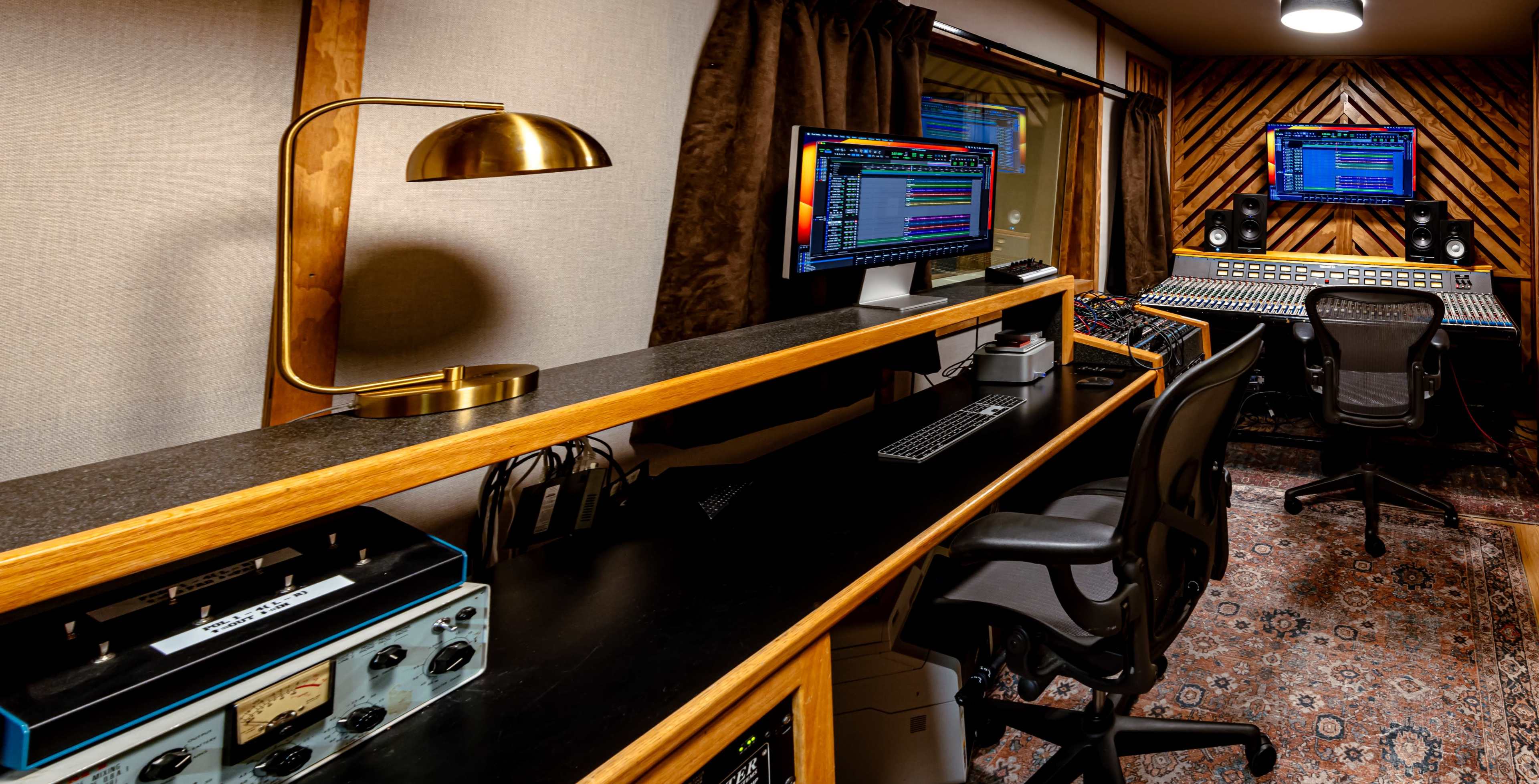 A recording studio featuring a large mixing console, dual computer monitors, and various audio equipment arranged on a wooden counter.