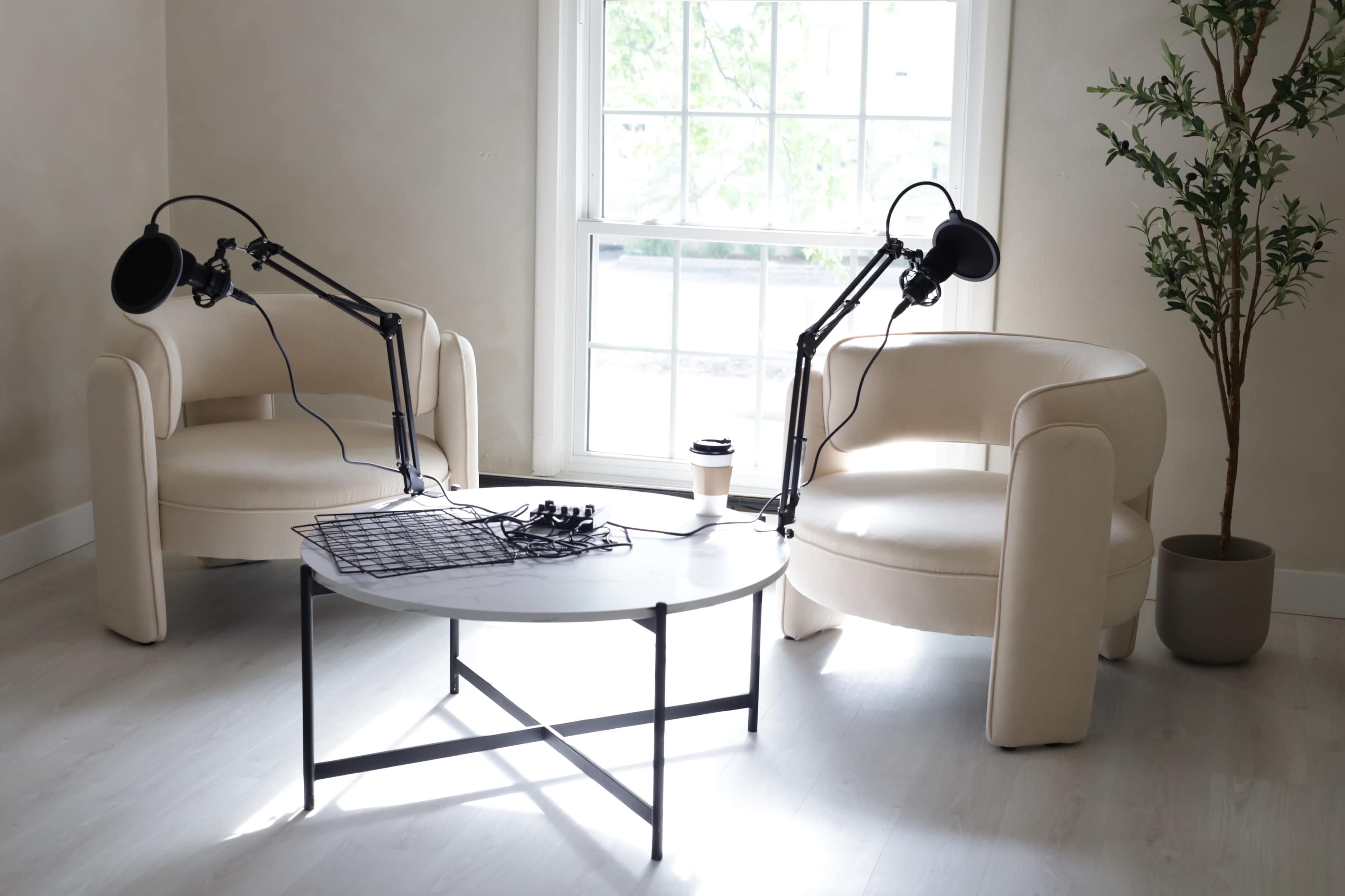 Two cream-colored chairs with adjustable microphone stands beside a round white coffee table in a well-lit room with a large window.