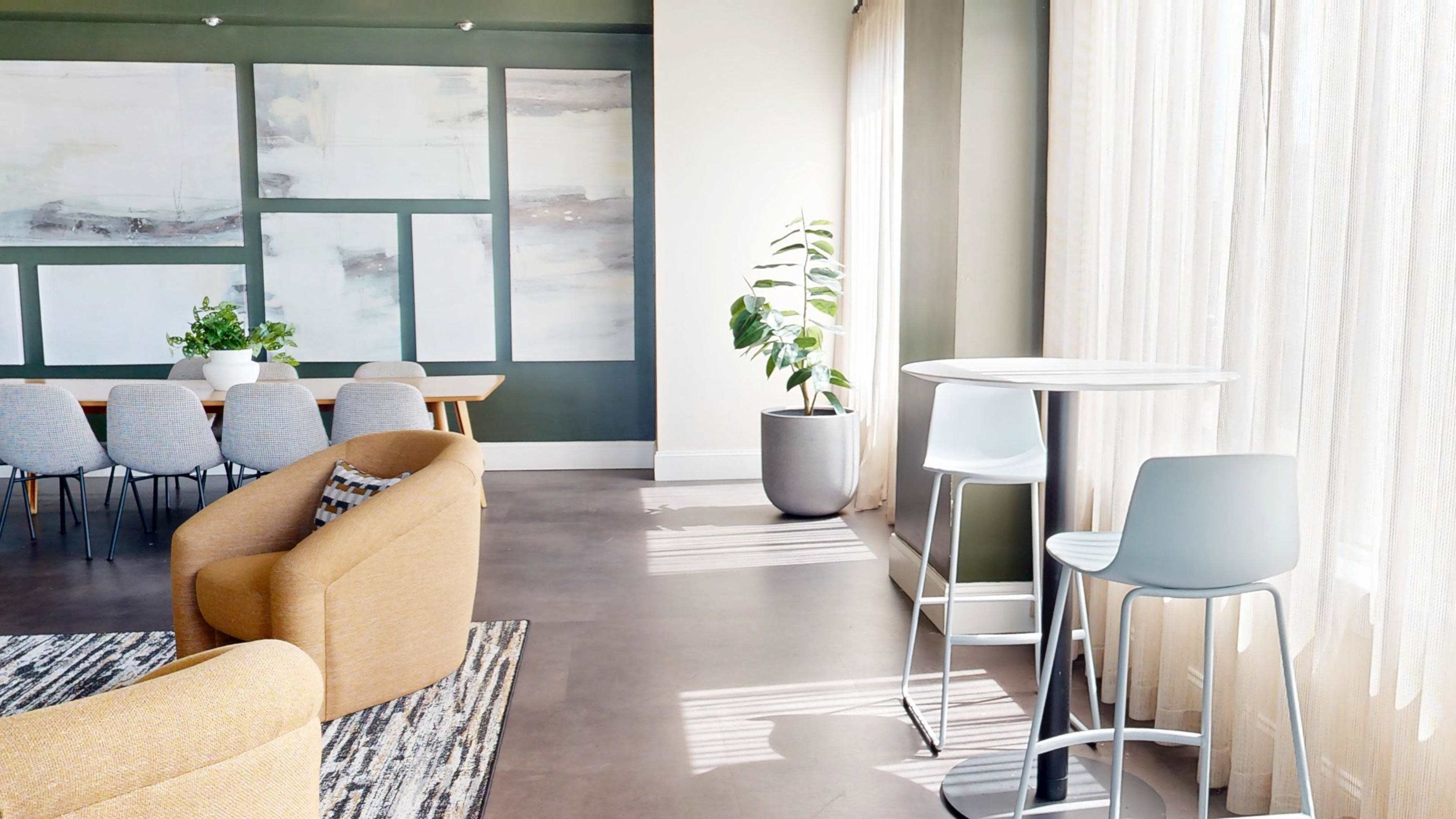 The image shows a modern interior space featuring a dining area with a long table and chairs, a small round high table, a plant, and large windows with sheer curtains.