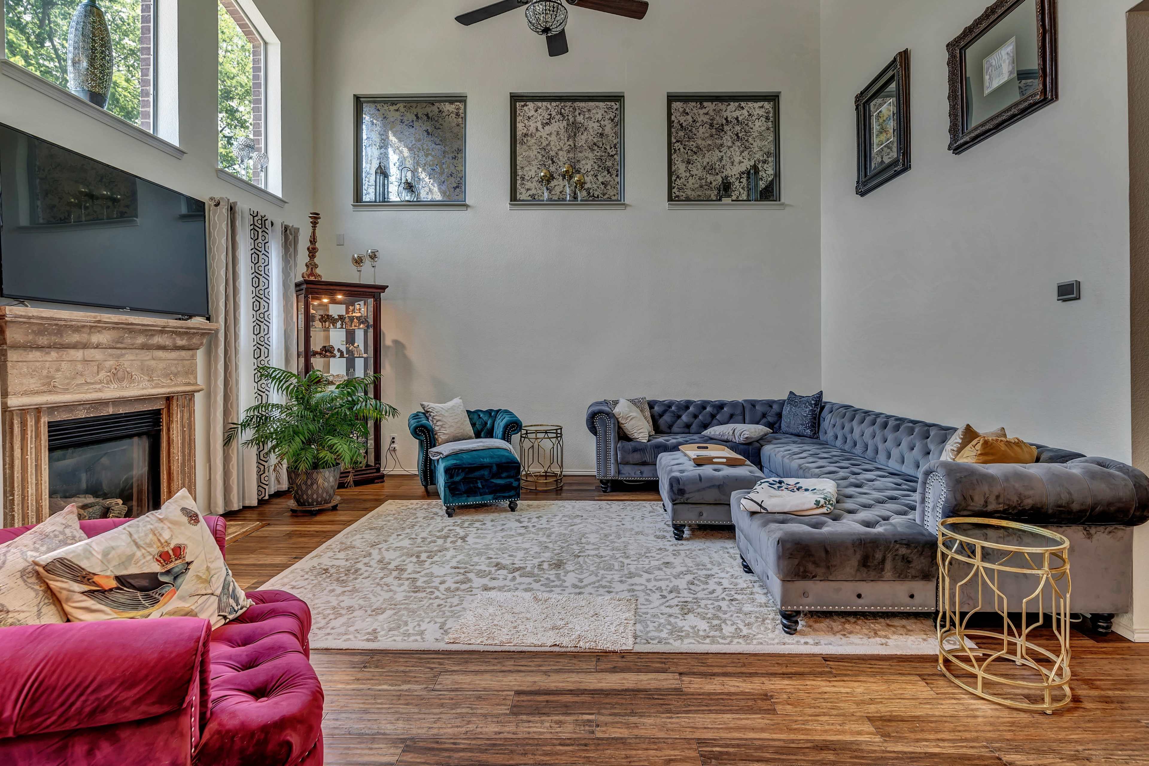 The living room features a sectional sofa, a plush armchair, and a decorative rug on wooden flooring, with large windows allowing natural light to enter.