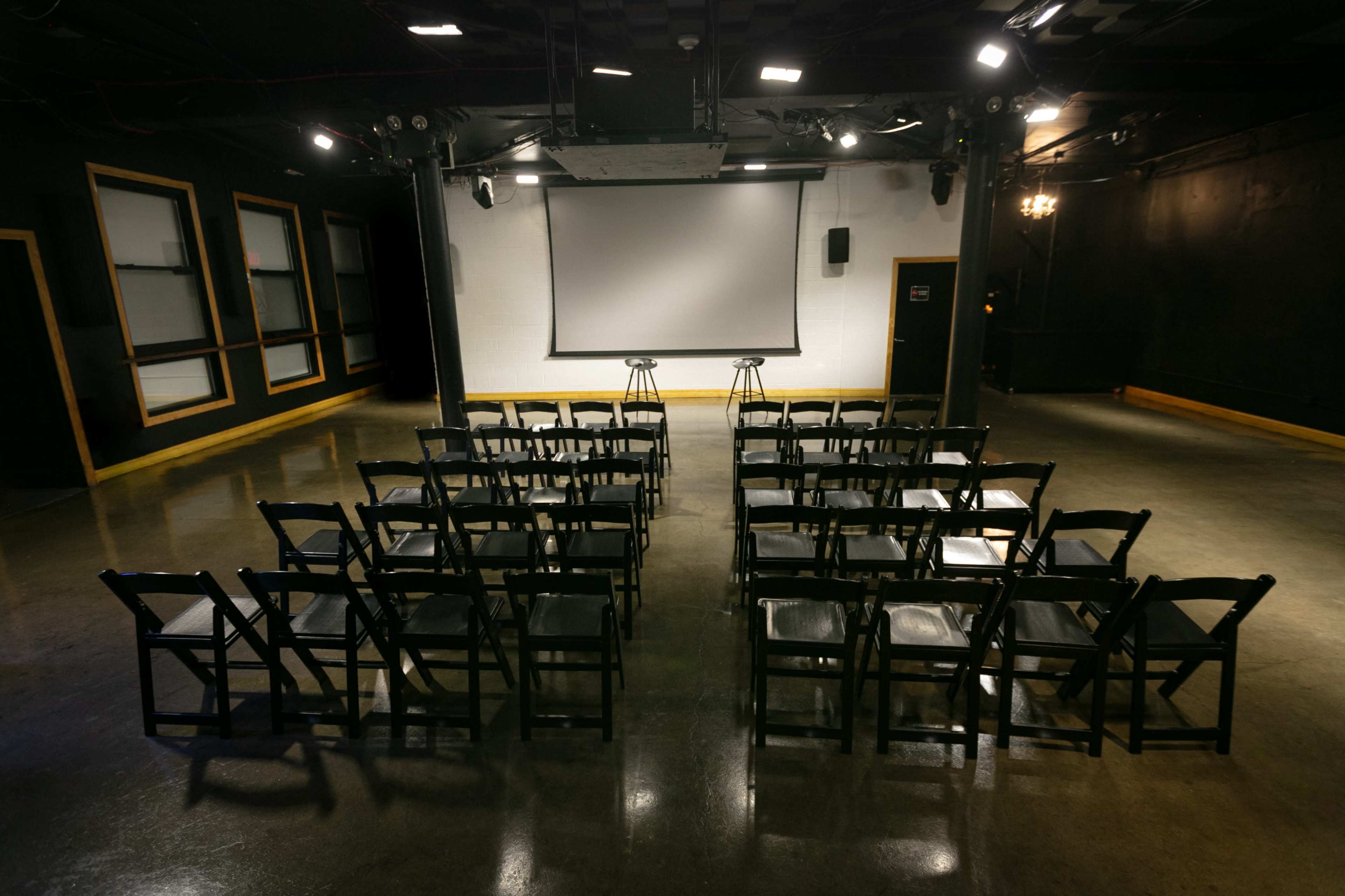 A presentation space is set up with rows of black chairs facing a screen and two stools.