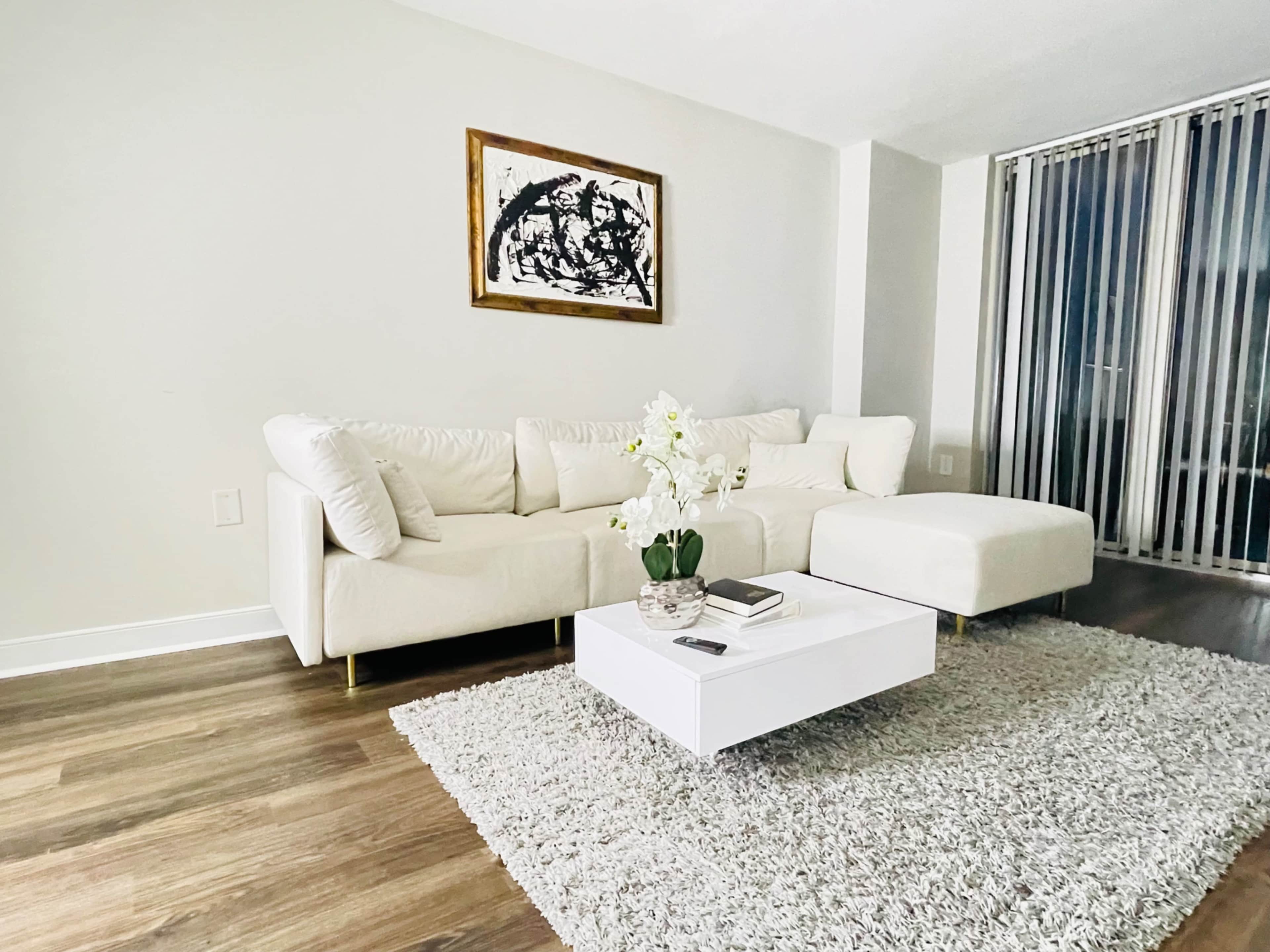 A modern living room with a white sectional sofa, a low coffee table, and a large area rug on wooden flooring, complemented by a piece of artwork on the wall and tall windows.
