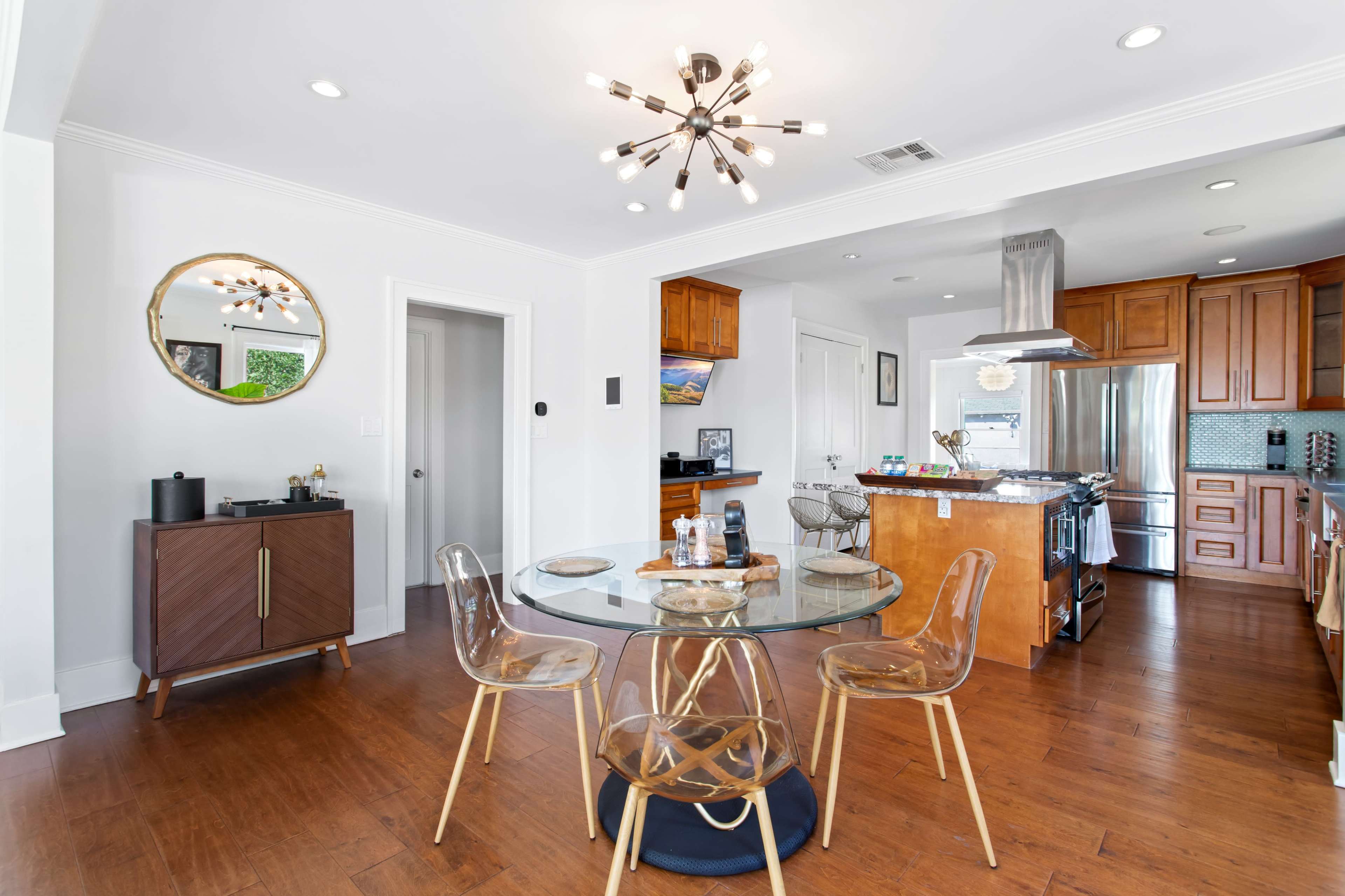 A modern kitchen and dining area featuring a round glass dining table with transparent chairs, a wooden cabinet, and a kitchen with wooden cabinetry and stainless steel appliances.
