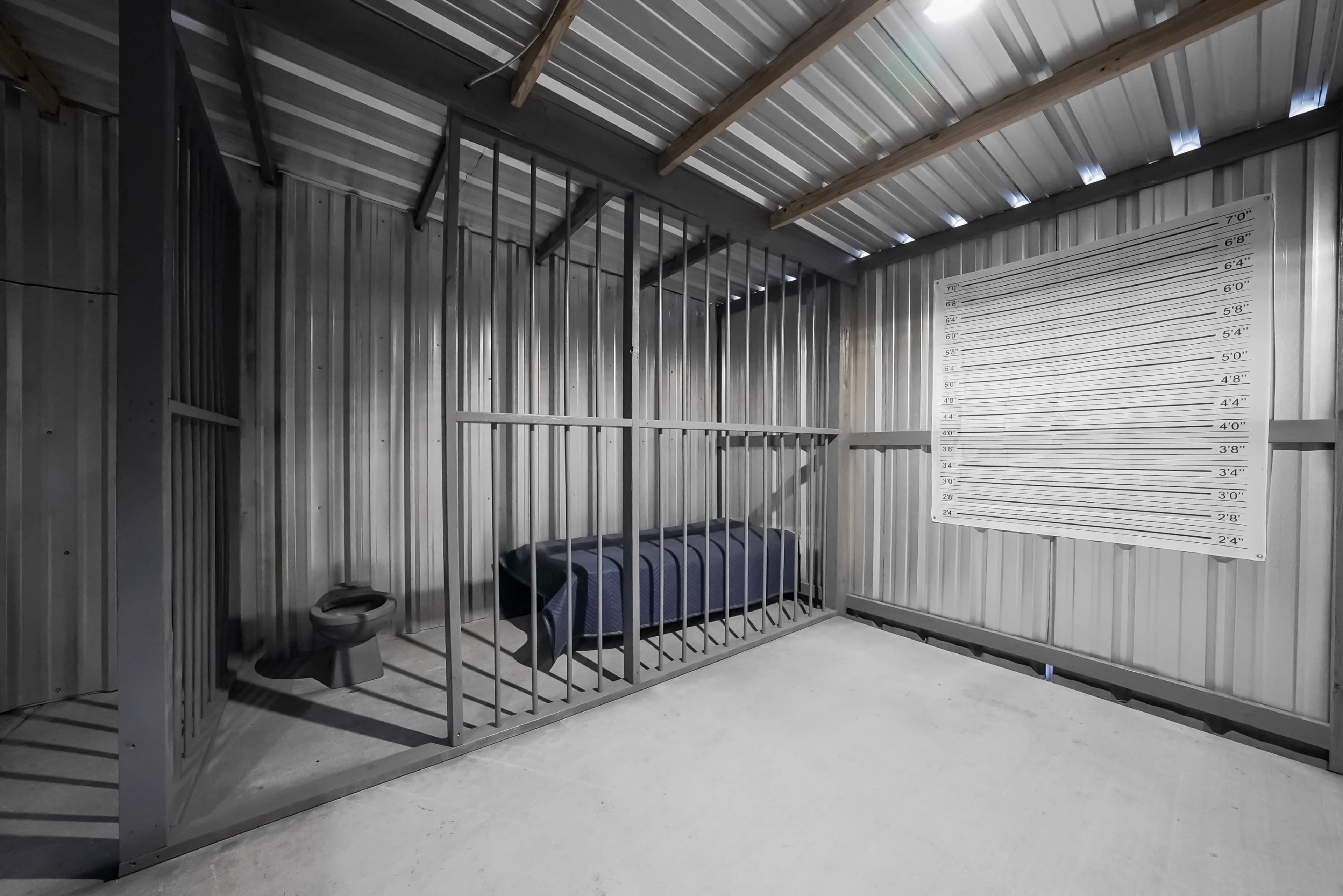 The image shows a stark, industrial room with metal bars on one side, a bed covered with a dark blanket, and a height chart on the adjacent wall.