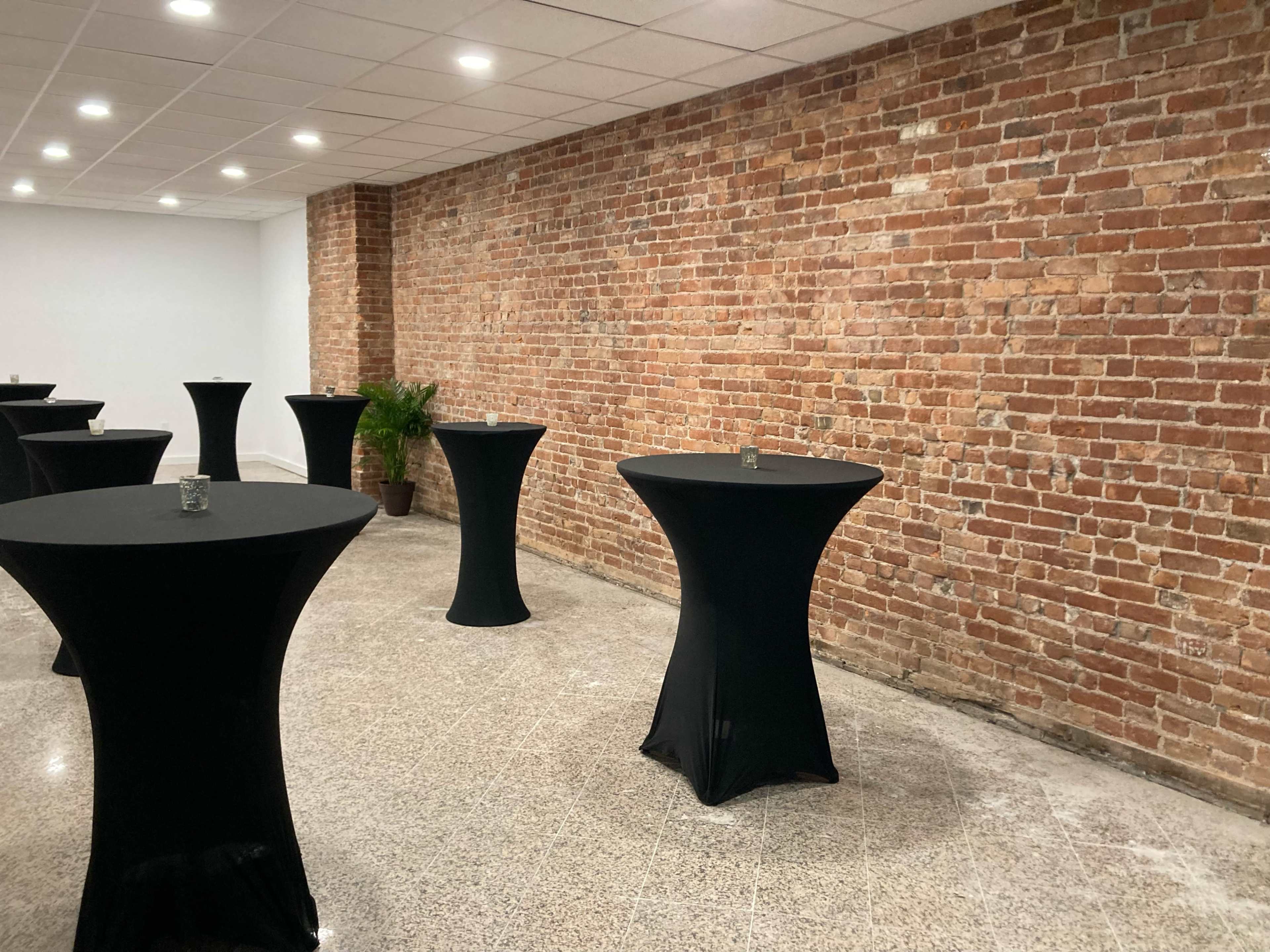 A spacious room with several tall black cocktail tables set against a brick wall.