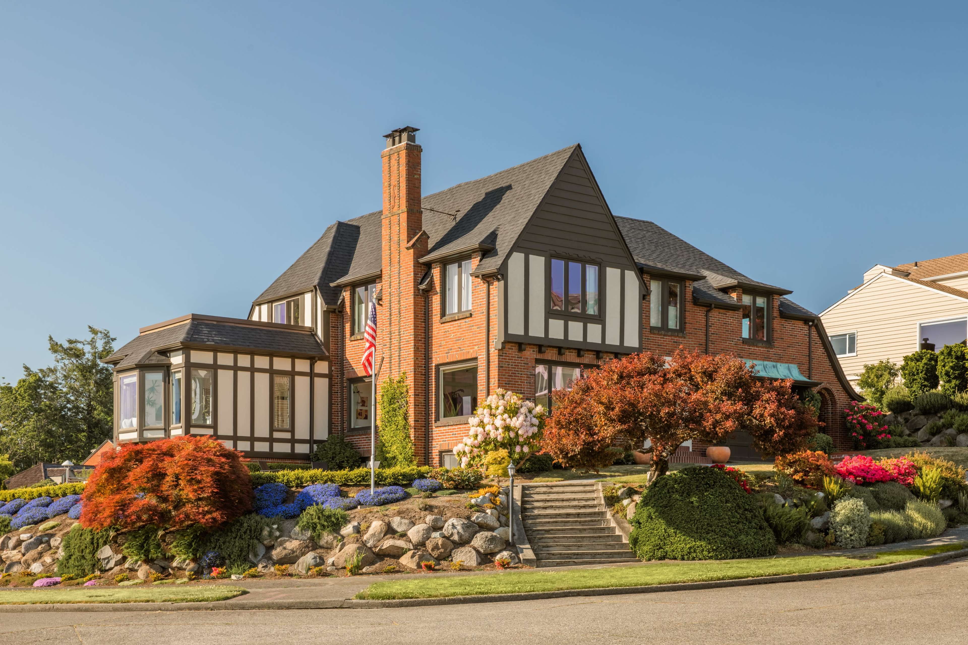 A large, two-story brick house with a steep roof, surrounded by well-maintained landscaping and colorful flowerbeds.