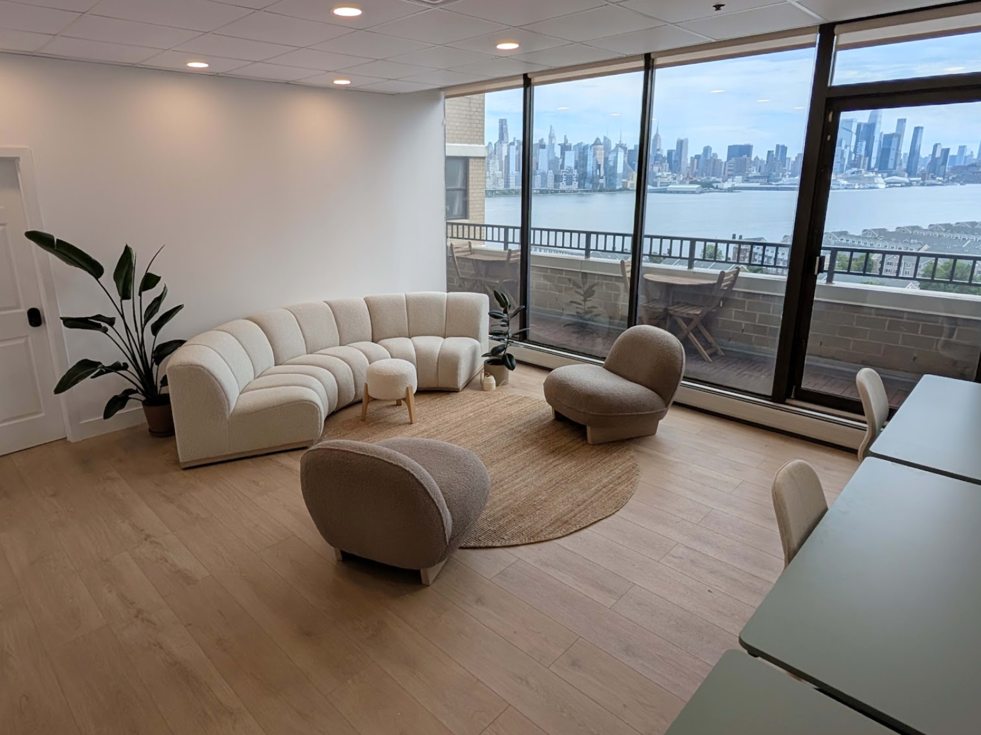 The image shows a modern, minimalist living room with a curved, light-colored sofa, a round rug, and a large window displaying a view of a city skyline and waterfront.