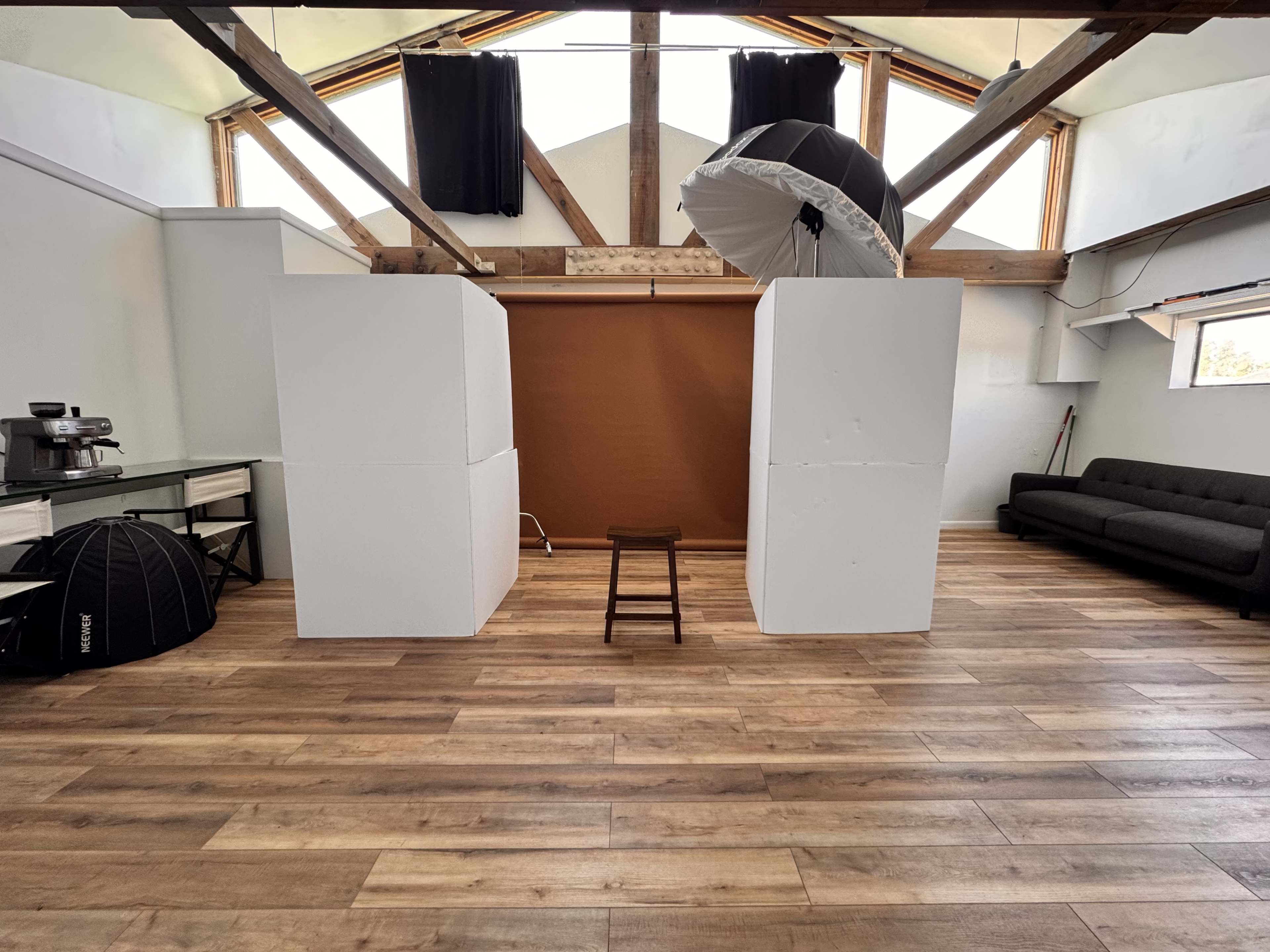 The image shows a spacious studio space with two large white backdrops and a brown backdrop in the center, a stool in front, and a sofa against the wall.