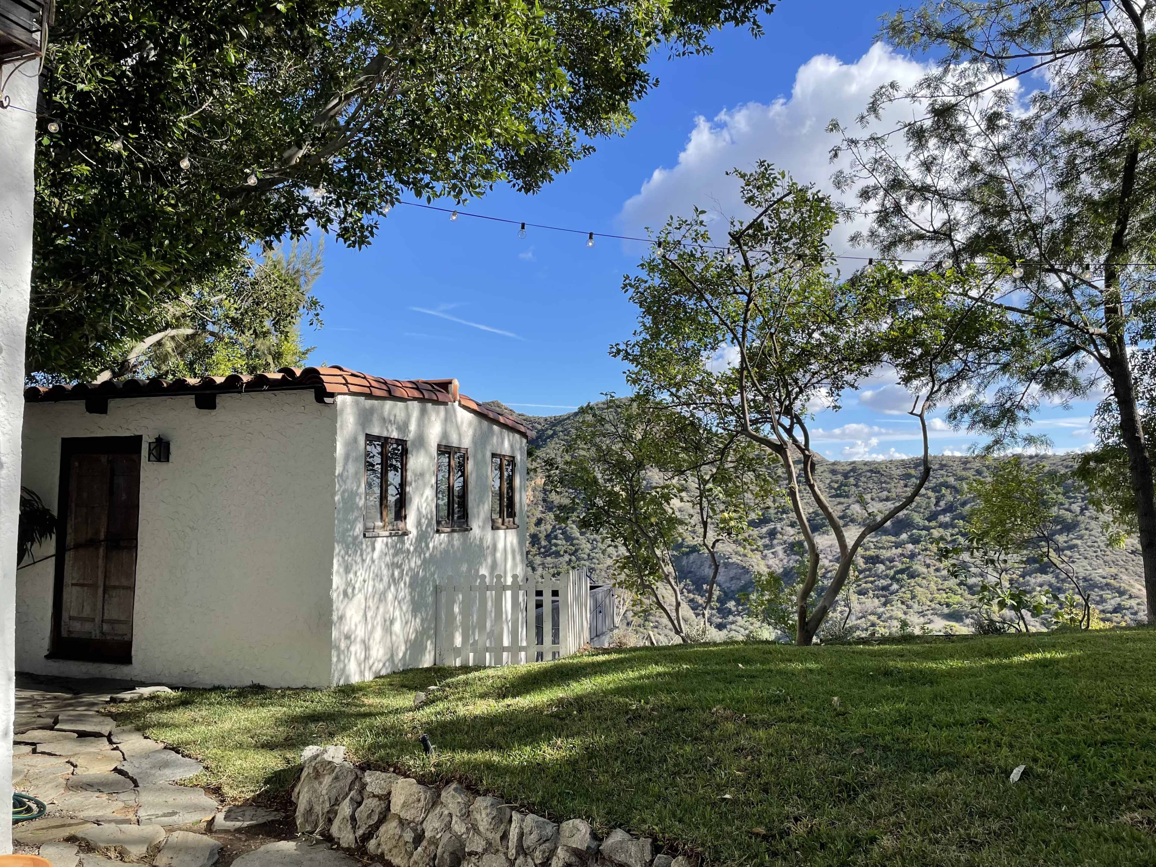 A white stucco house with a tile roof sits beside a grassy area, surrounded by trees and mountains in the background.