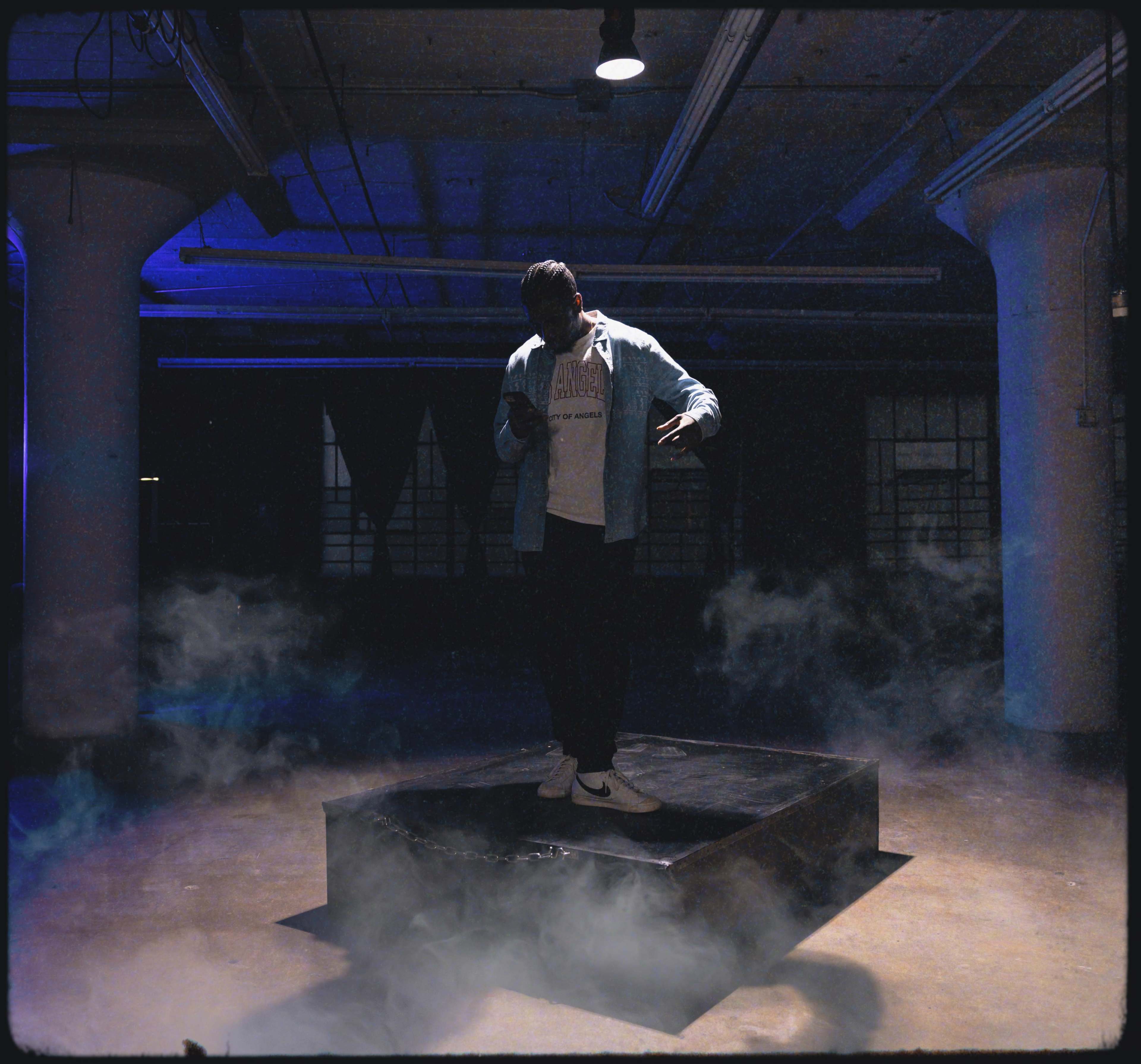 A person stands on a raised platform in a dimly lit industrial space, surrounded by wisps of smoke.