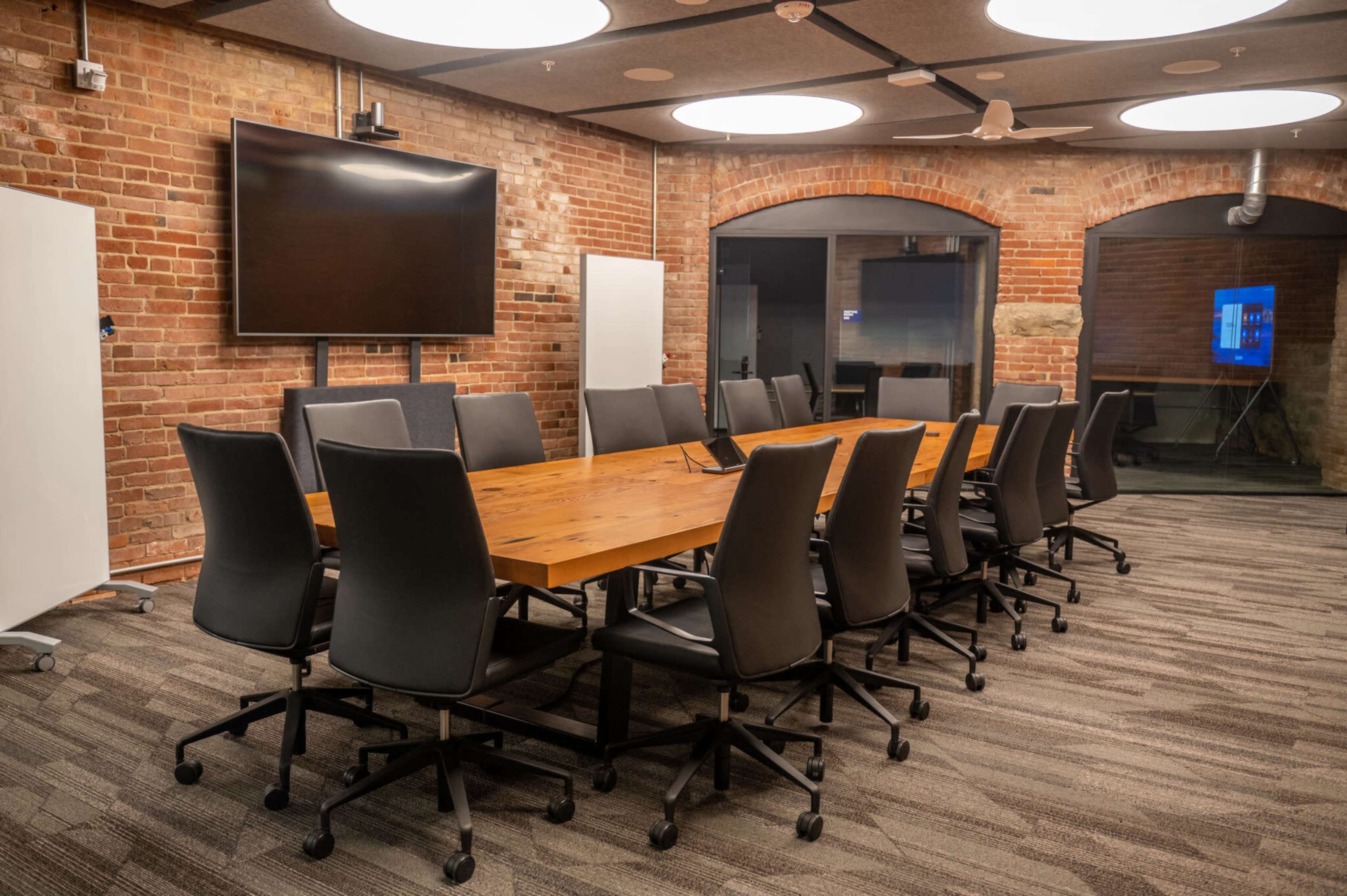 A large conference room features a long wooden table surrounded by gray office chairs, with a large screen mounted on the brick wall.