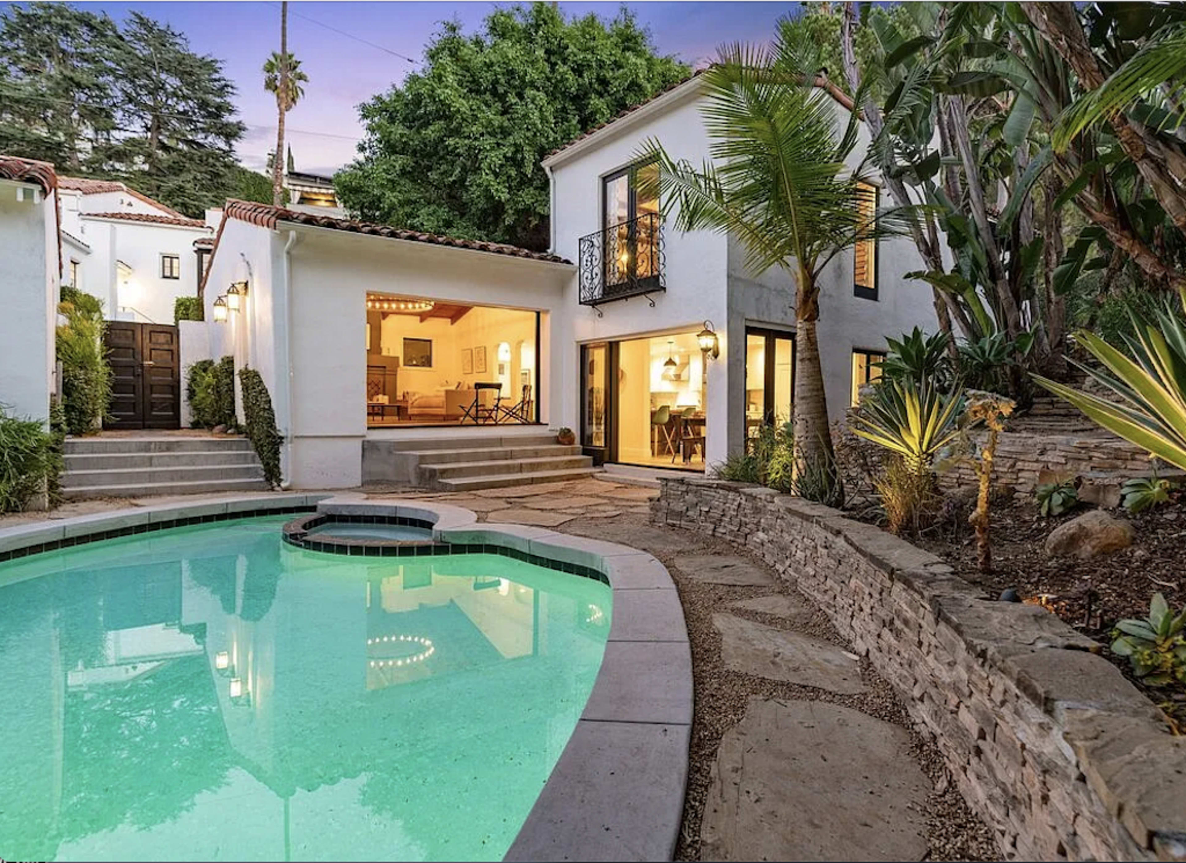 The image shows a private pool surrounded by stone pathways and tropical plants, with a two-story house featuring large windows and an open living area visible in the background.