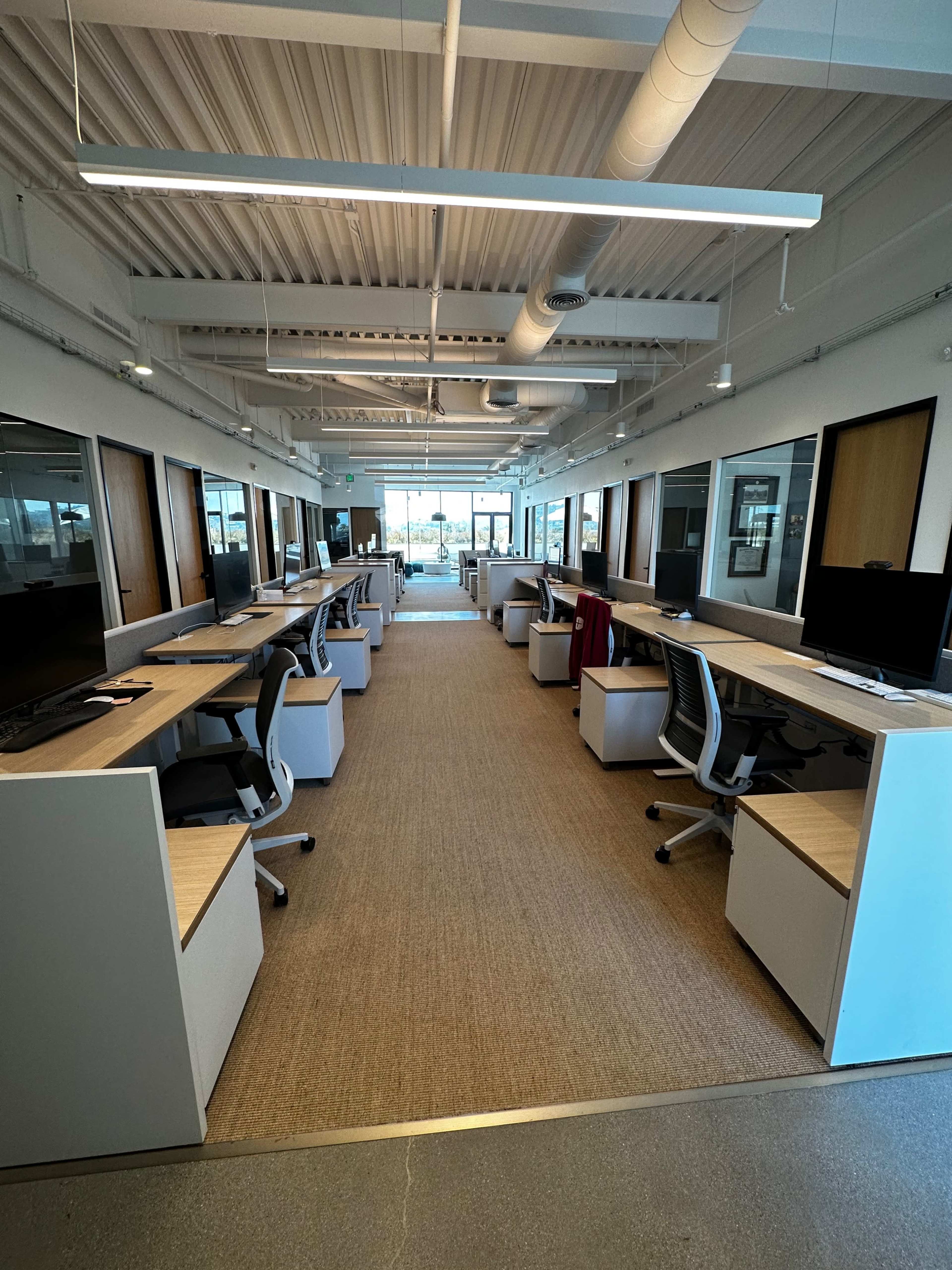 The image shows a modern office space with rows of desks, each equipped with a computer and ergonomic chairs, along a central hallway leading to glass doors.