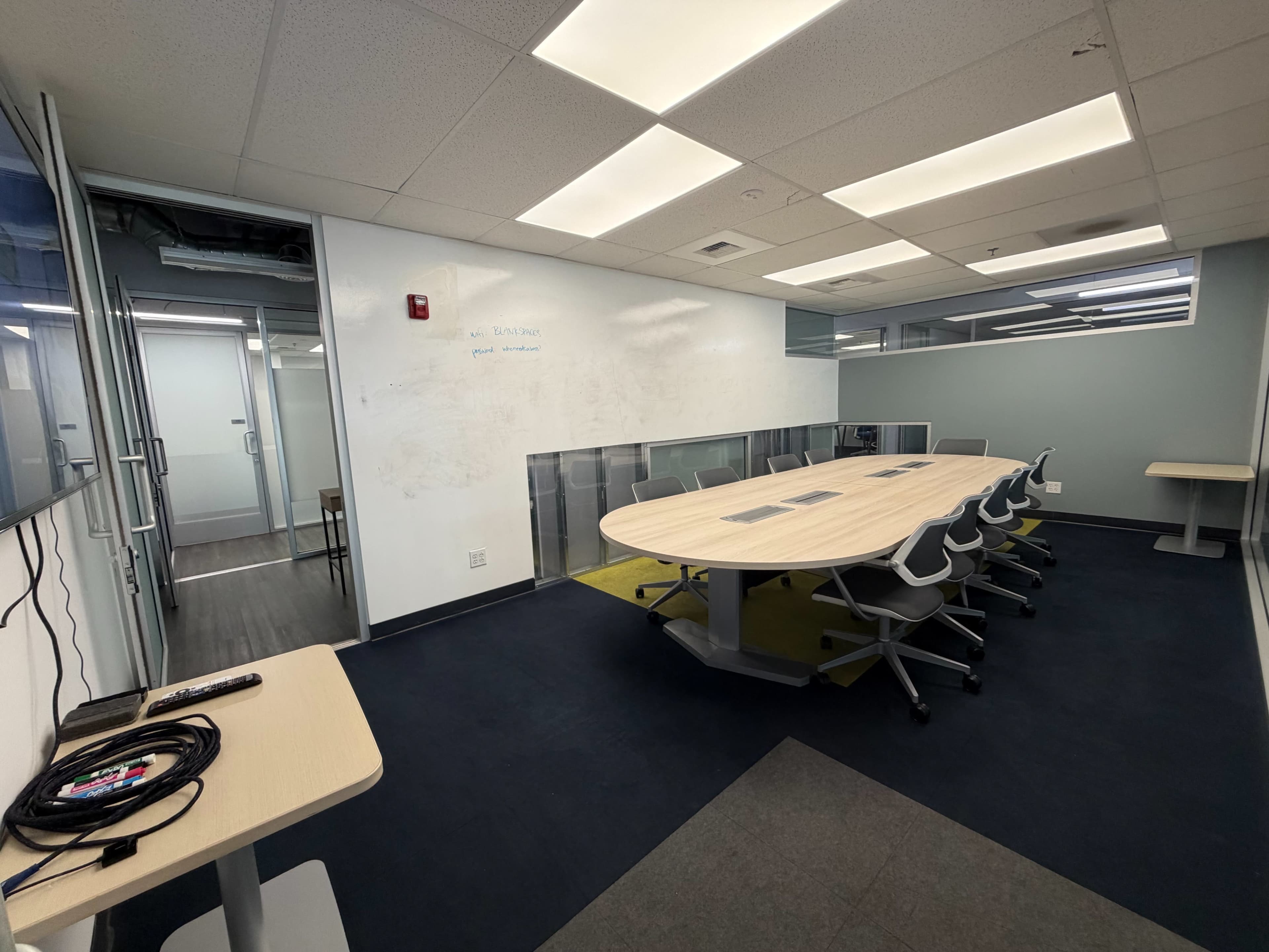 The image shows a modern conference room with a long table surrounded by ergonomic chairs, glass walls, and a digital display on one side.