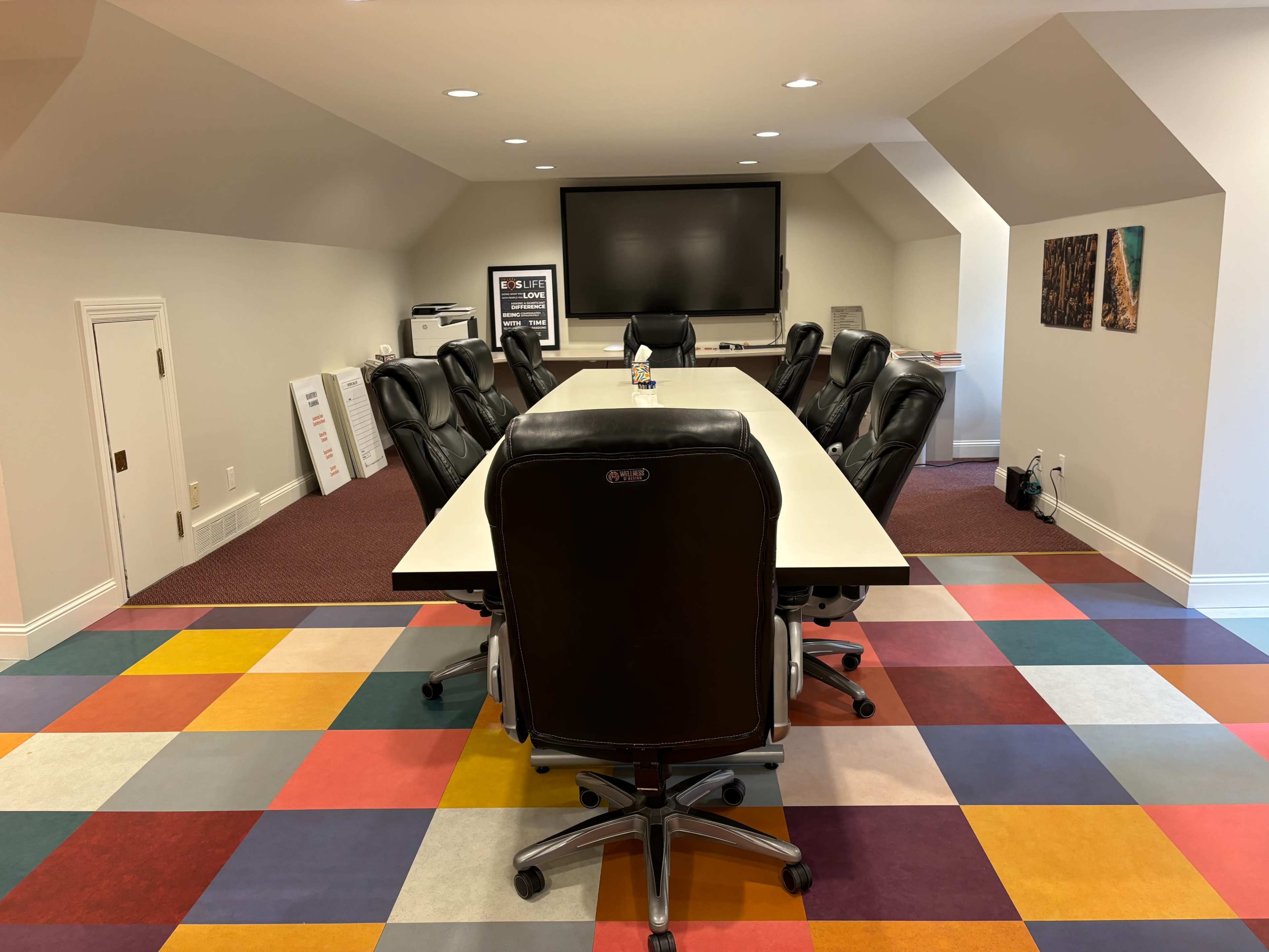 A conference room featuring a large rectangular table surrounded by black office chairs, with a television screen mounted on the wall and colorful patterned flooring.