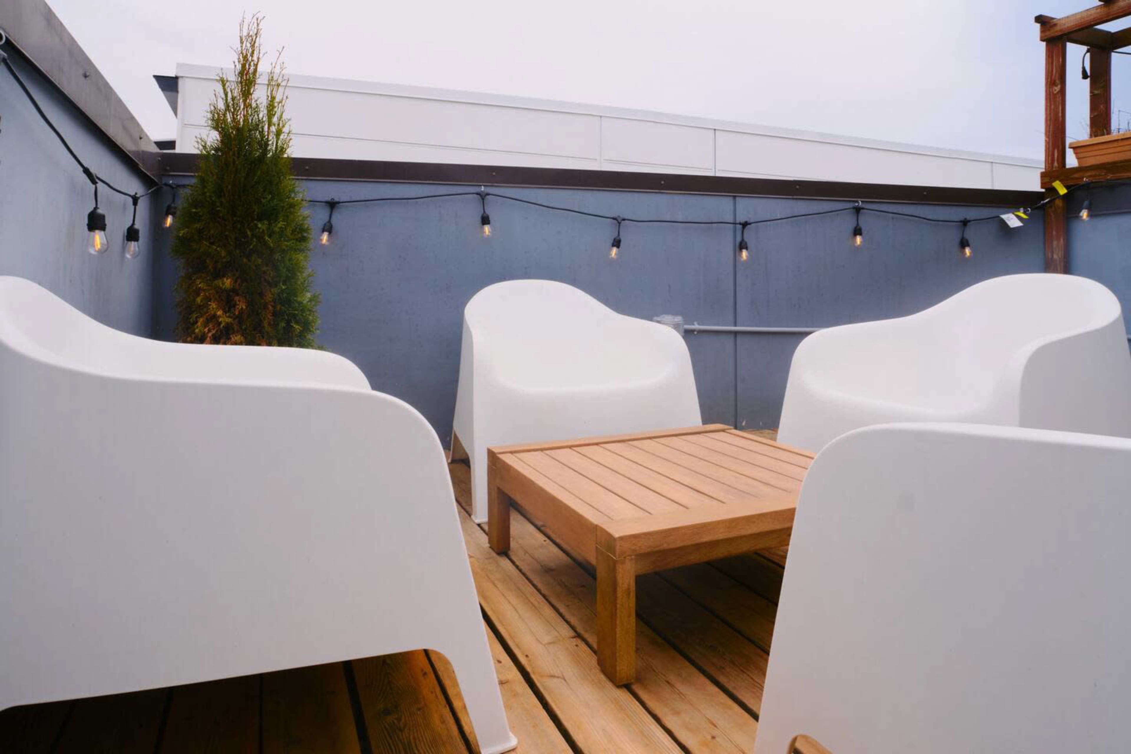 A rooftop patio features four white chairs surrounding a wooden table, with string lights overhead and a potted plant in the corner.