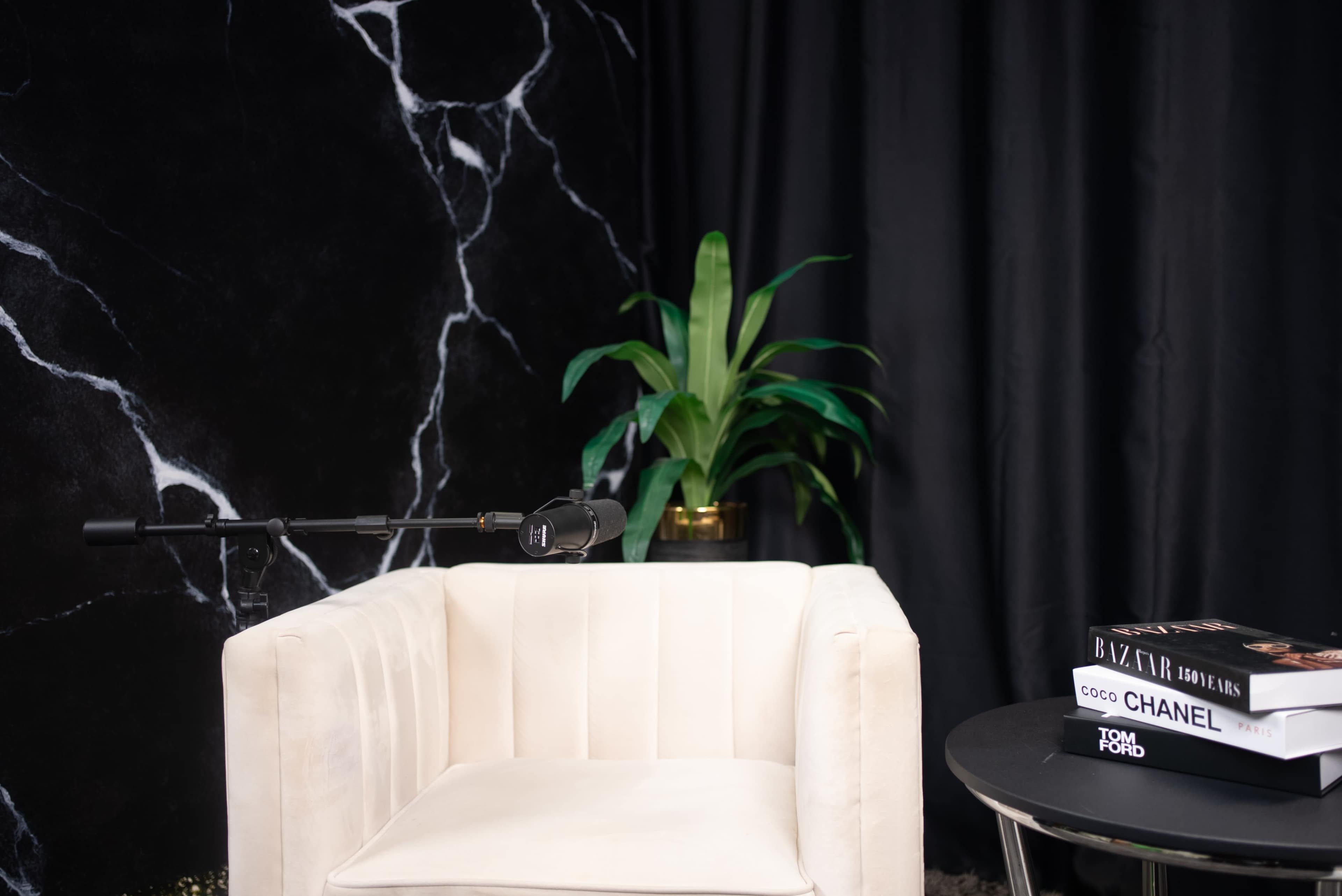 The image shows a modern, minimalist interview setup featuring a white armchair, a small black table with books, a microphone on a stand, and a green plant against a black, marbled backdrop.