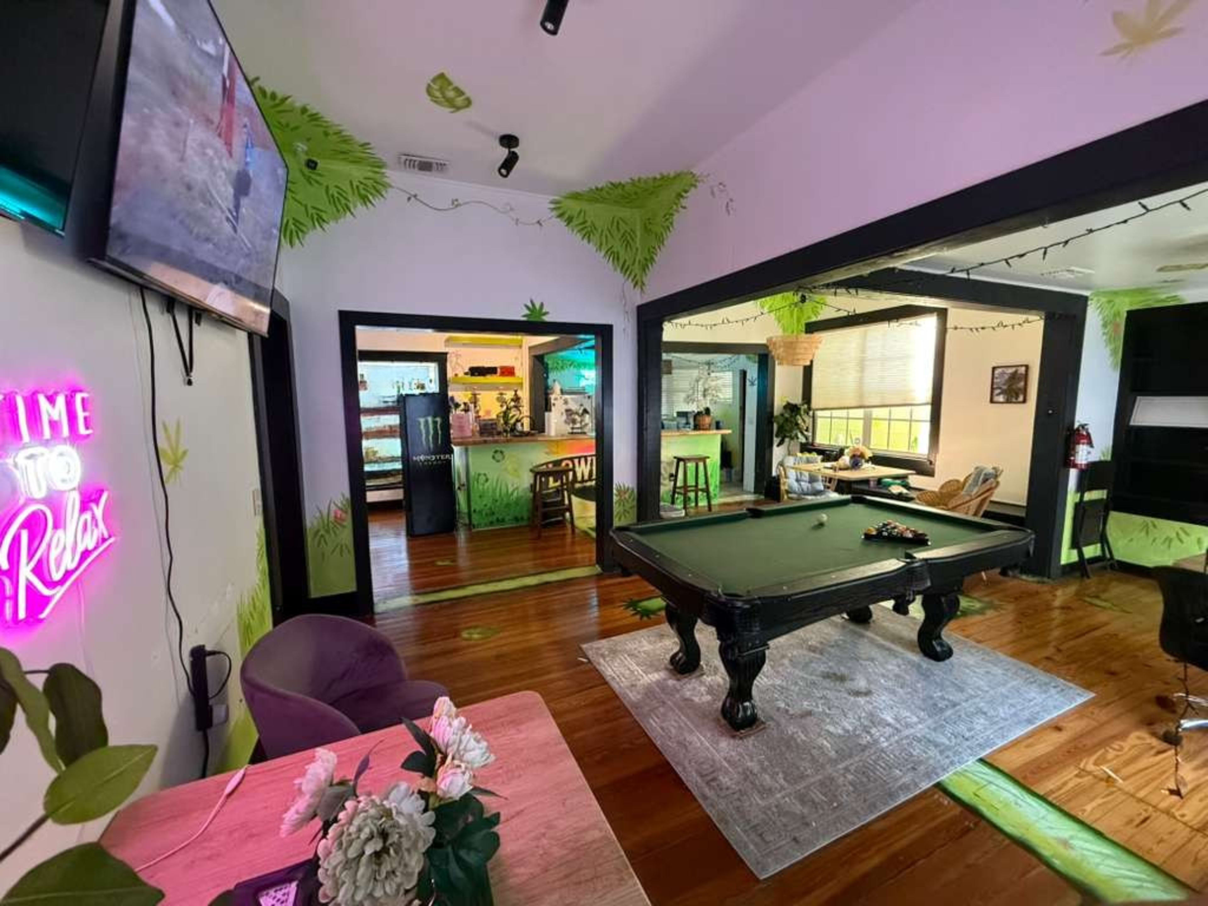 The image shows a recreational room featuring a pool table, greenery-themed decor, a neon sign, and a view into a kitchen area.