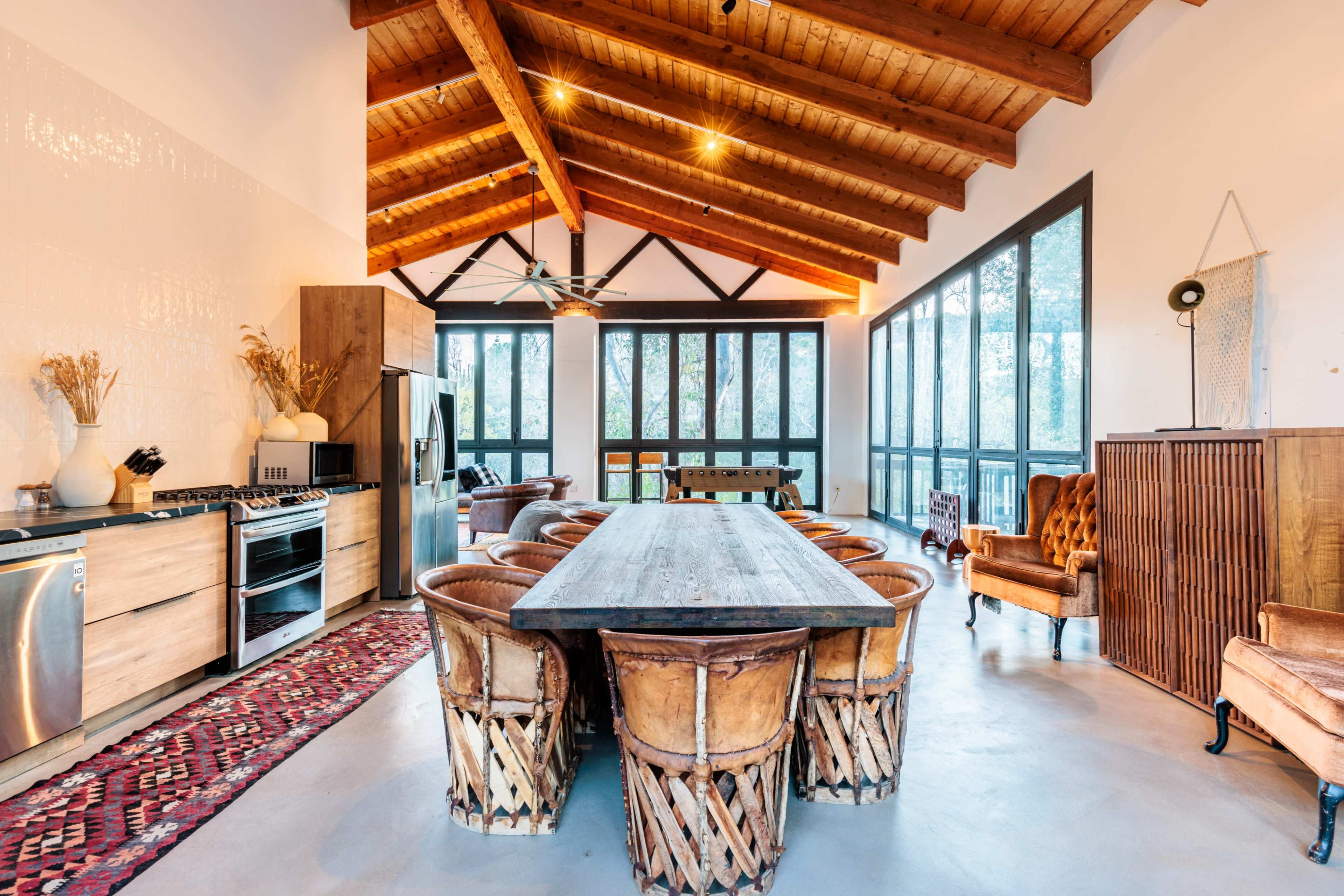 A spacious dining room features a large wooden table surrounded by woven chairs, with a high ceiling supported by exposed beams and large windows allowing for ample natural light.
