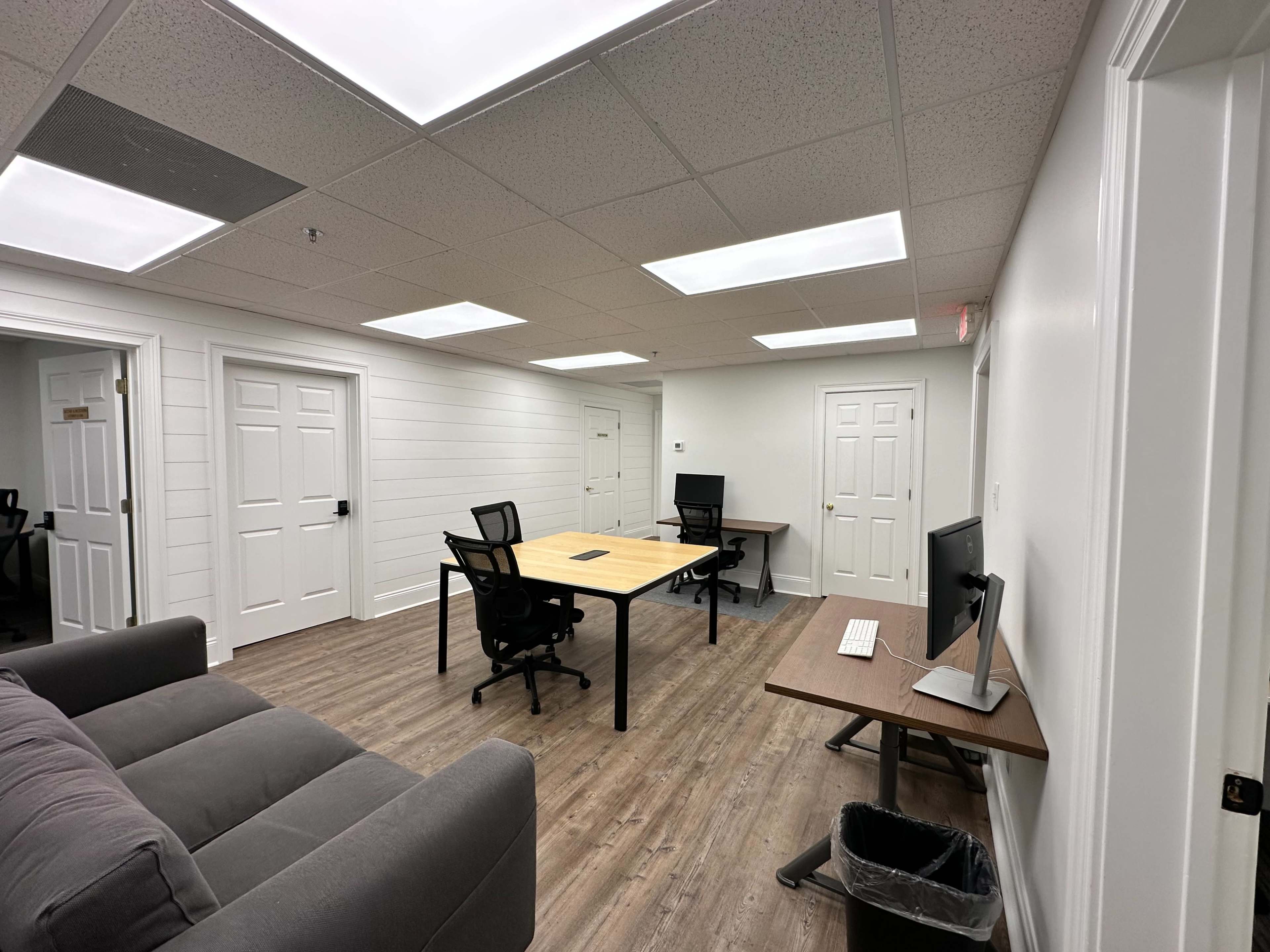 A modern, airy office space with two desks, a sofa, and bright overhead lights.