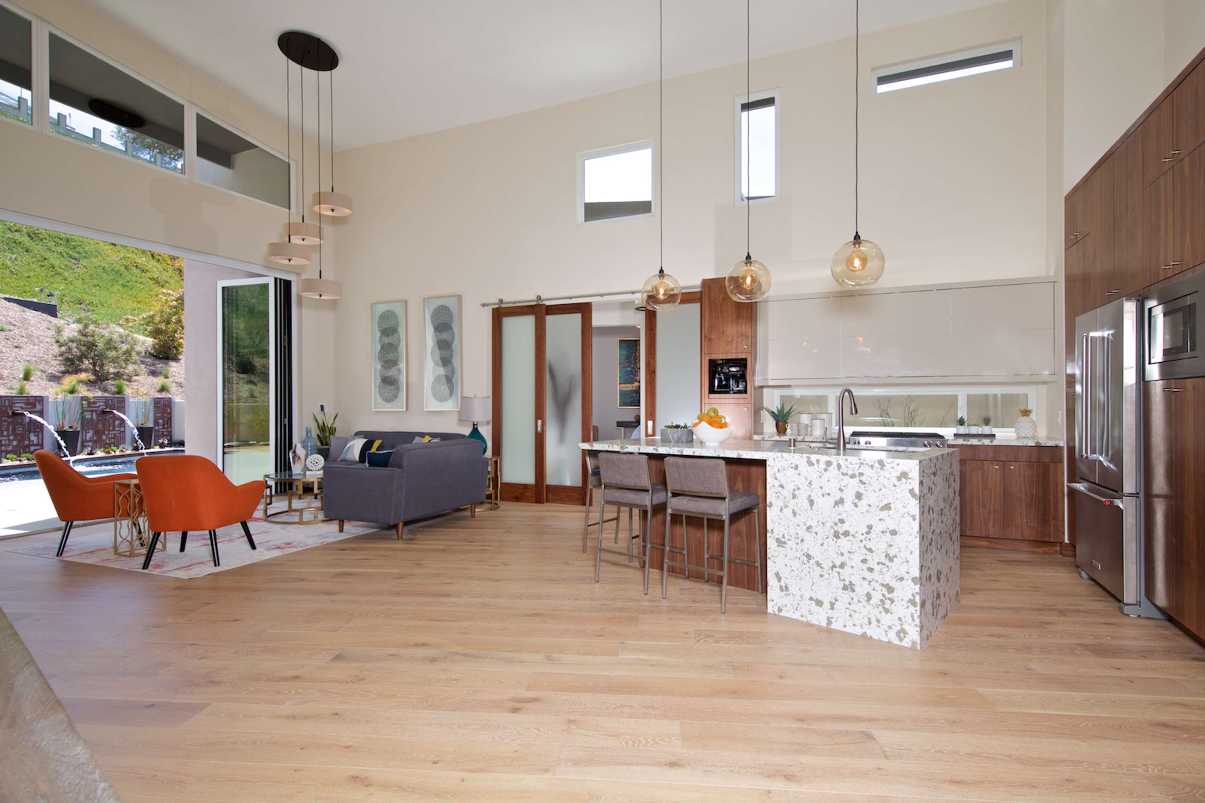 A modern open-concept kitchen and living area featuring a central island with a speckled countertop, contemporary furniture, and large windows leading to an outdoor space.
