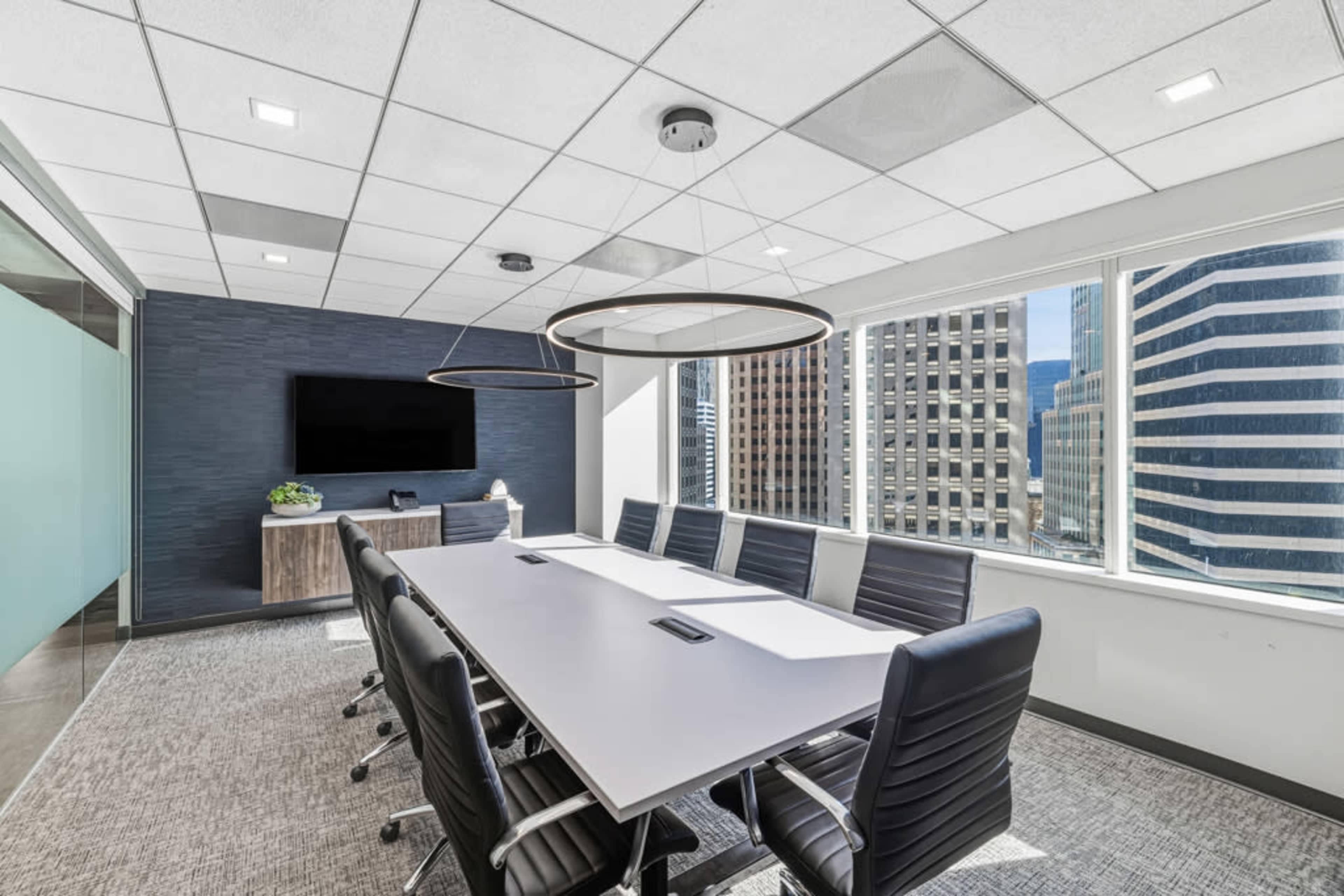 A modern conference room features a long table with ergonomic chairs, large windows offering a city view, and a wall-mounted screen.