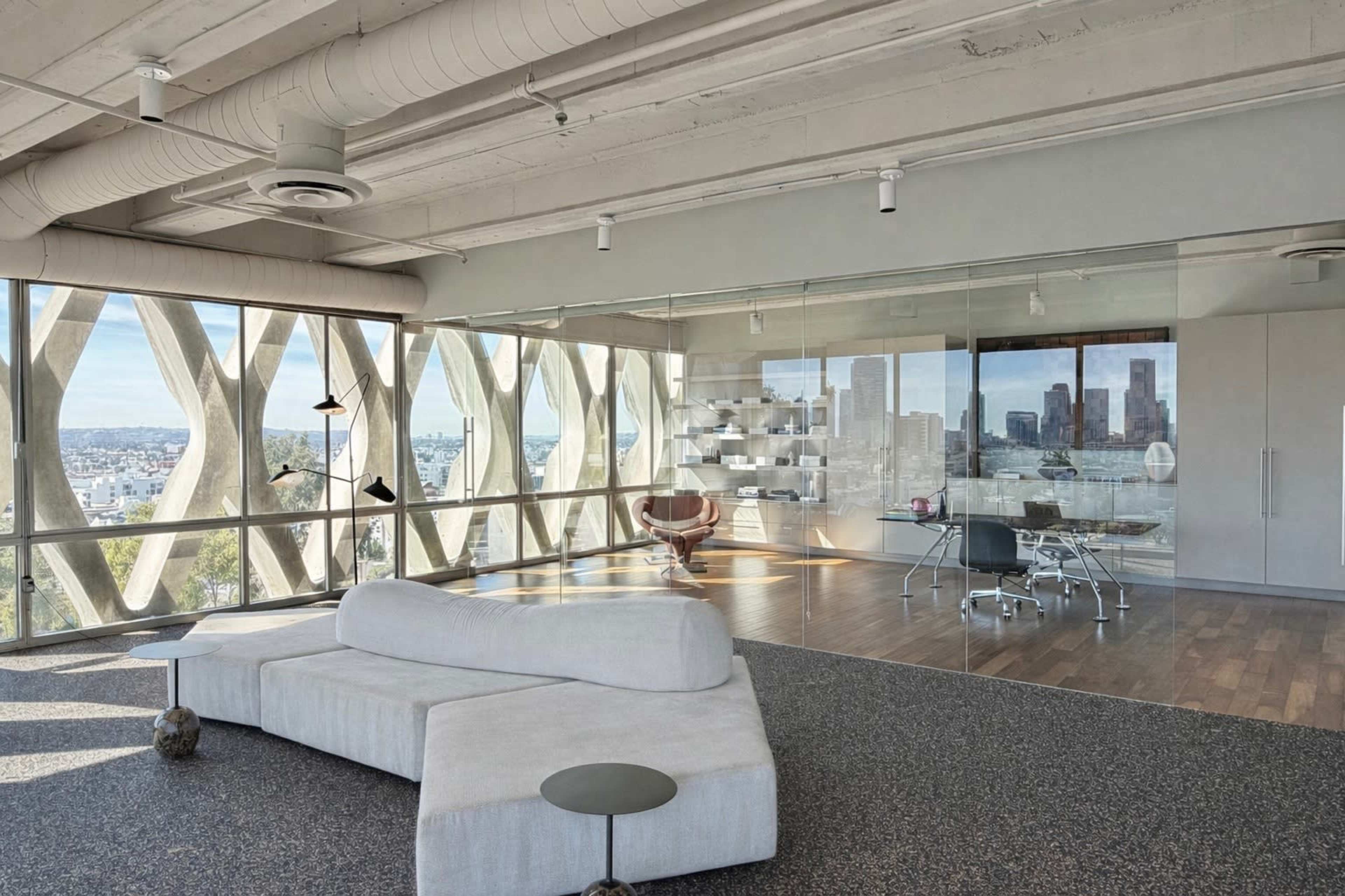 The image shows a modern open-concept living space with large windows offering a view of a city skyline, featuring a light gray sectional sofa and a glass-walled office area.