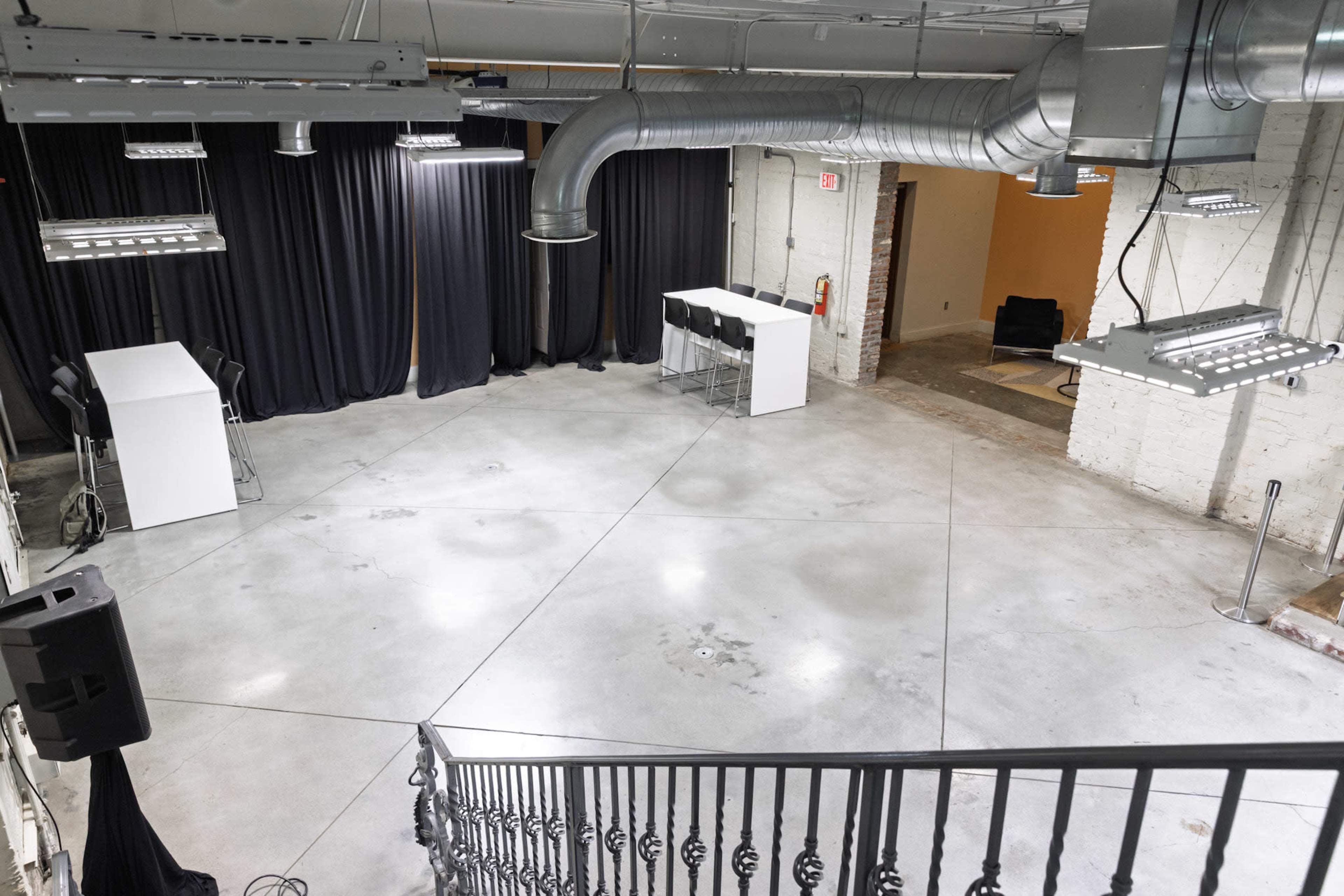 The image shows a spacious indoor venue with polished concrete flooring, dim lighting, a staircase, and black curtains in the background, along with two white tables at the front.