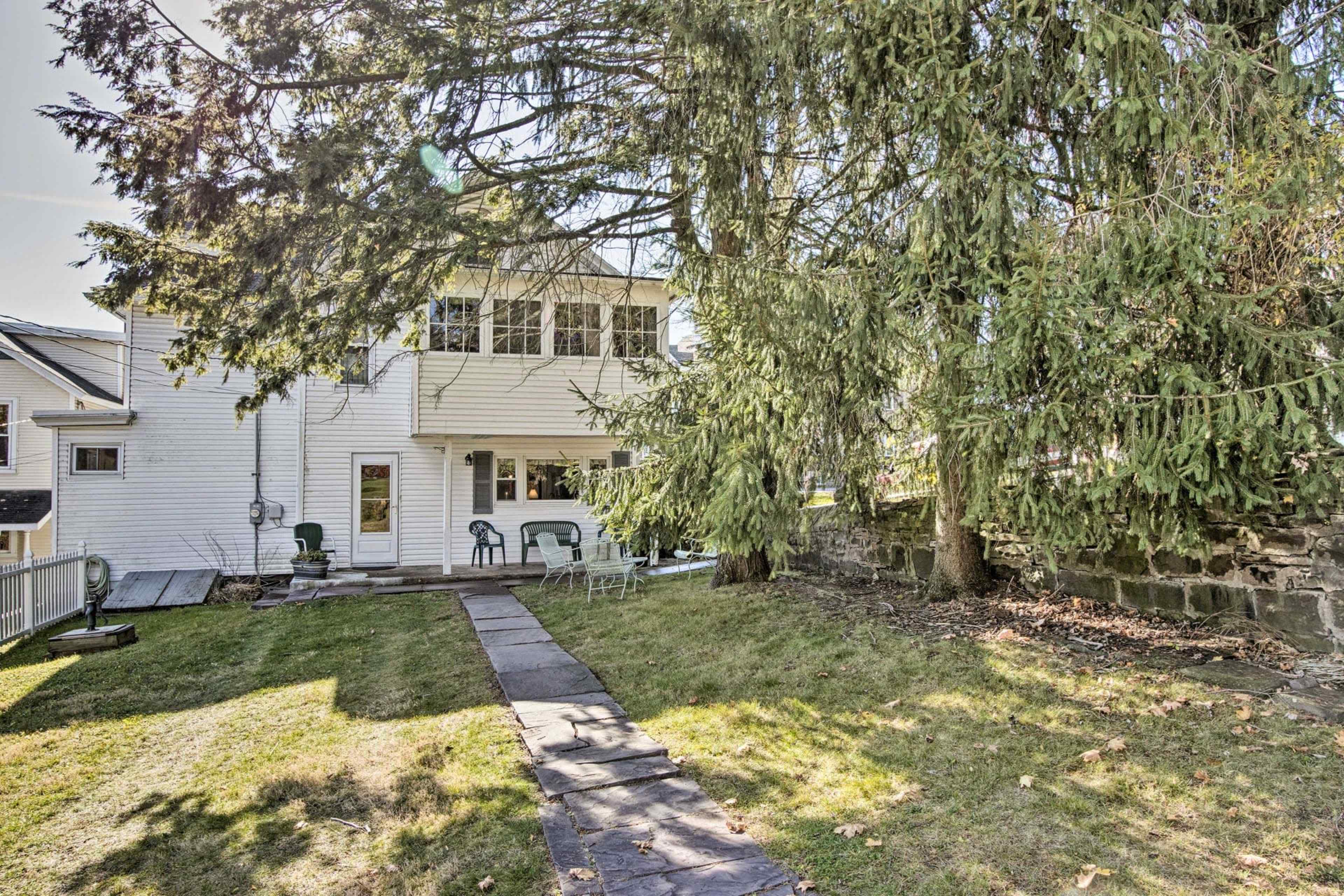 The image shows a two-story white house with a backyard featuring a path made of stones, a grassy area, and a large tree.