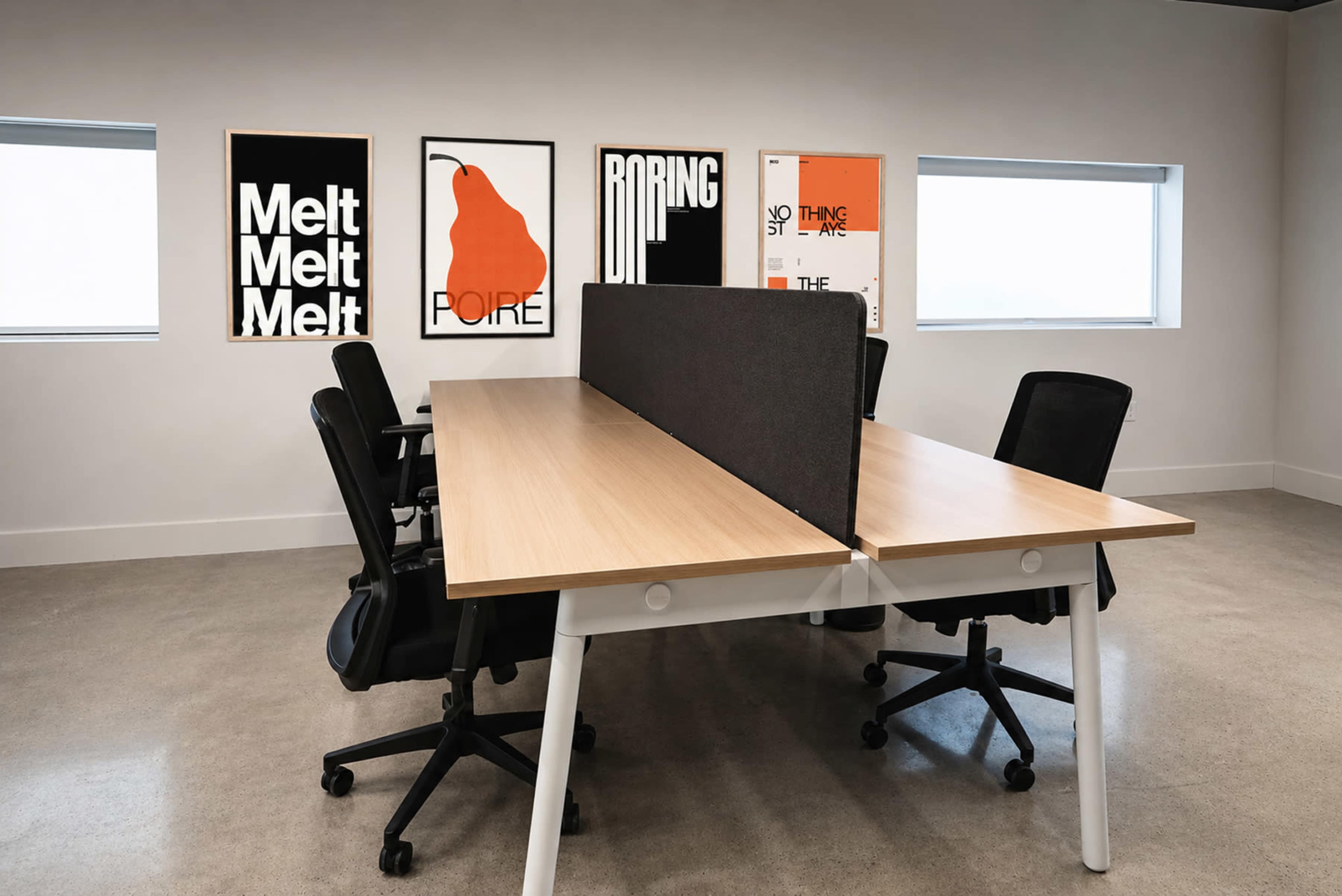 A modern office space features a long wooden desk divided by a gray partition, with black office chairs arranged around it and colorful artwork on the walls.