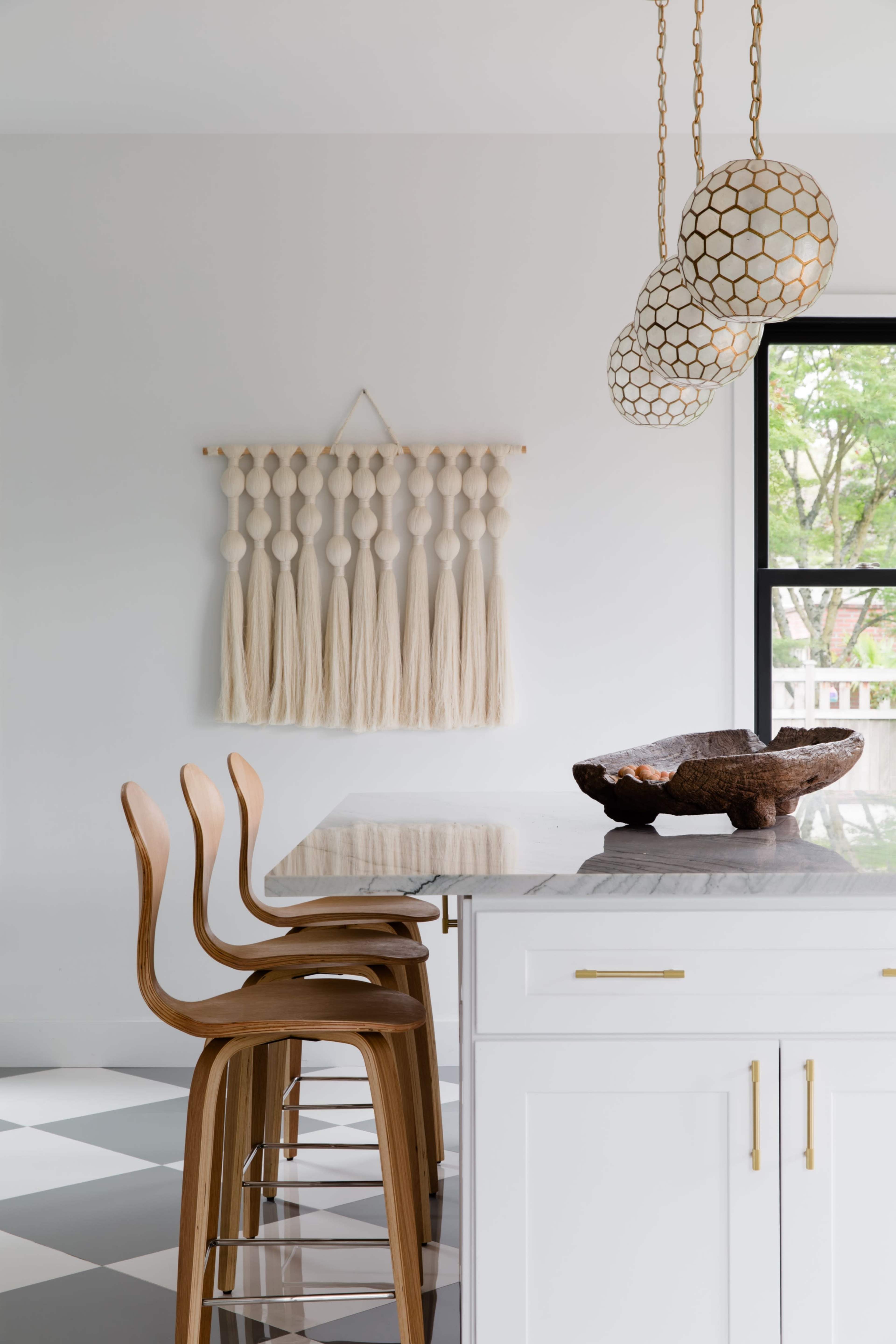 A modern kitchen features a marble countertop with wooden bar stools, a decorative wall hanging, and pendant lighting.
